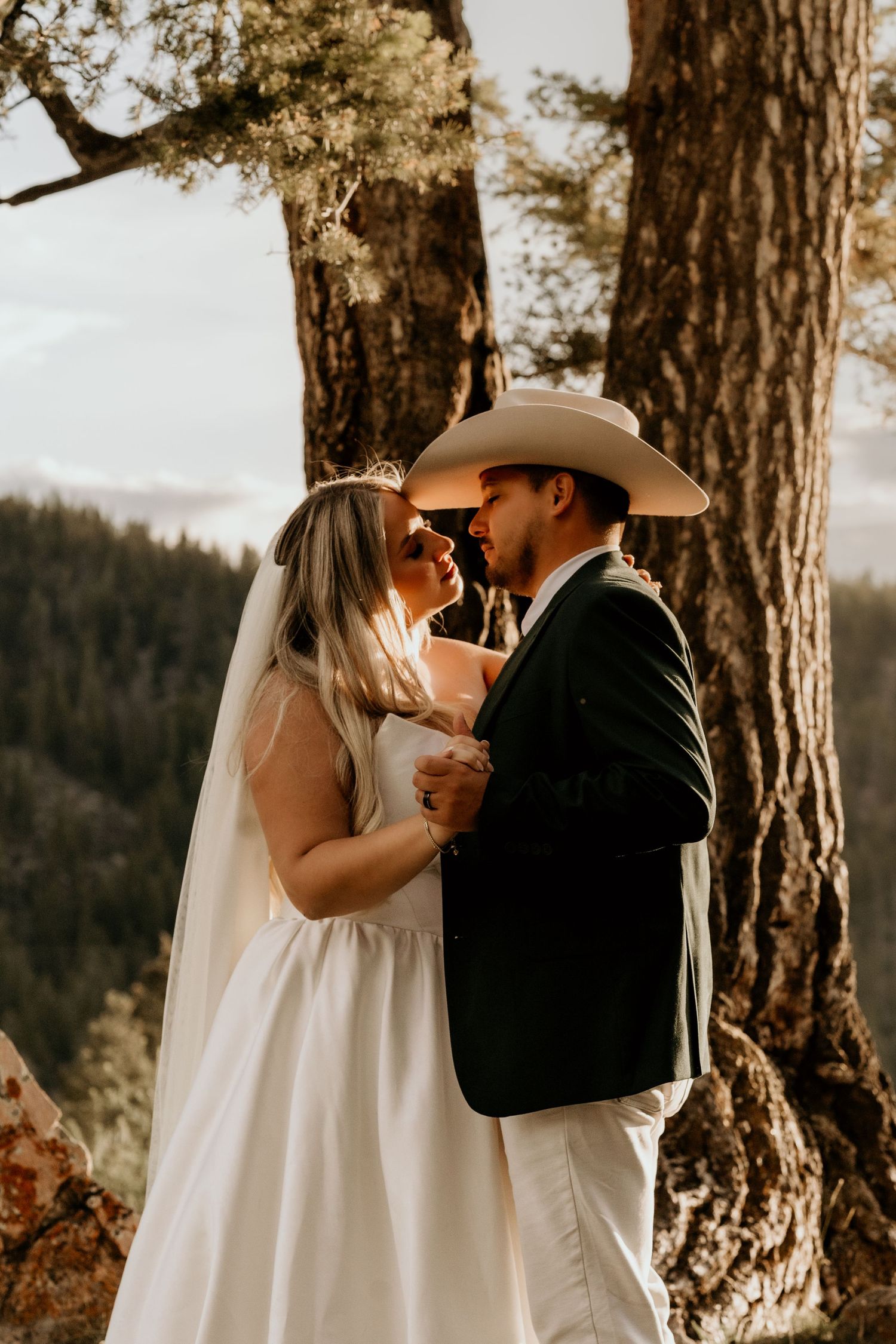 Bride and groom share a romantic moment under a pine tree with mountain views at a rustic outdoor wedding.