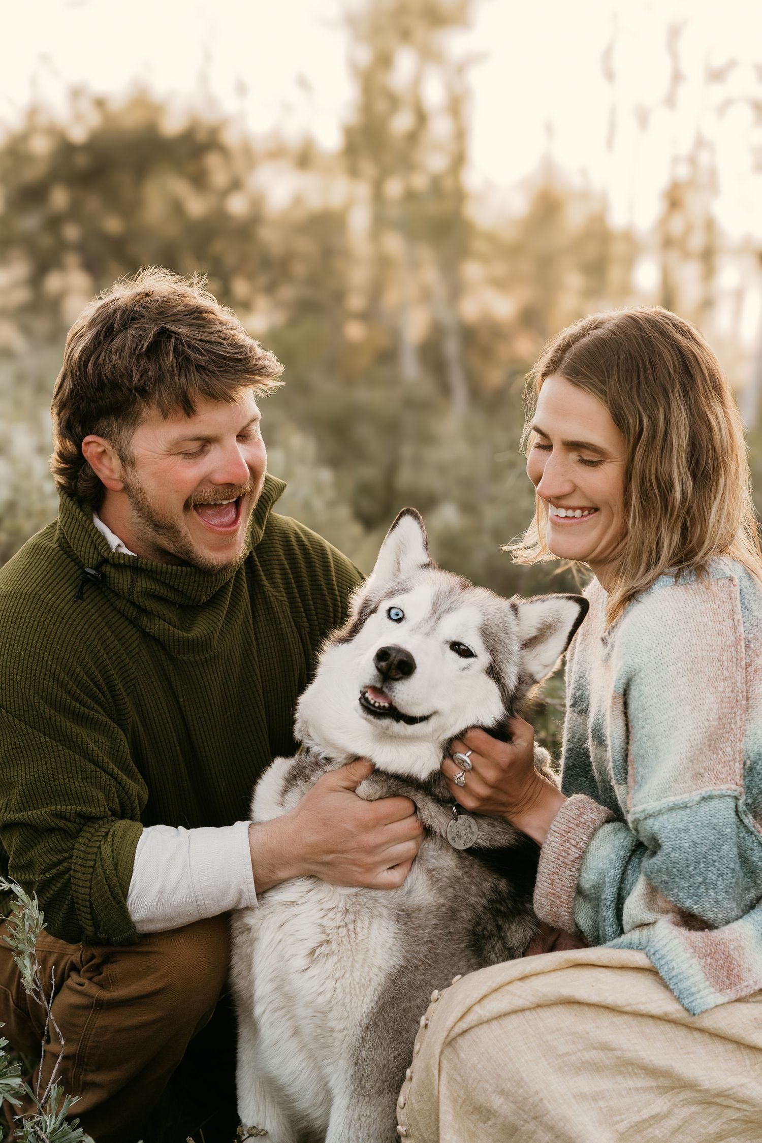 Smiling couple petting their Siberian Husky dog outdoors in warm autumn light.
