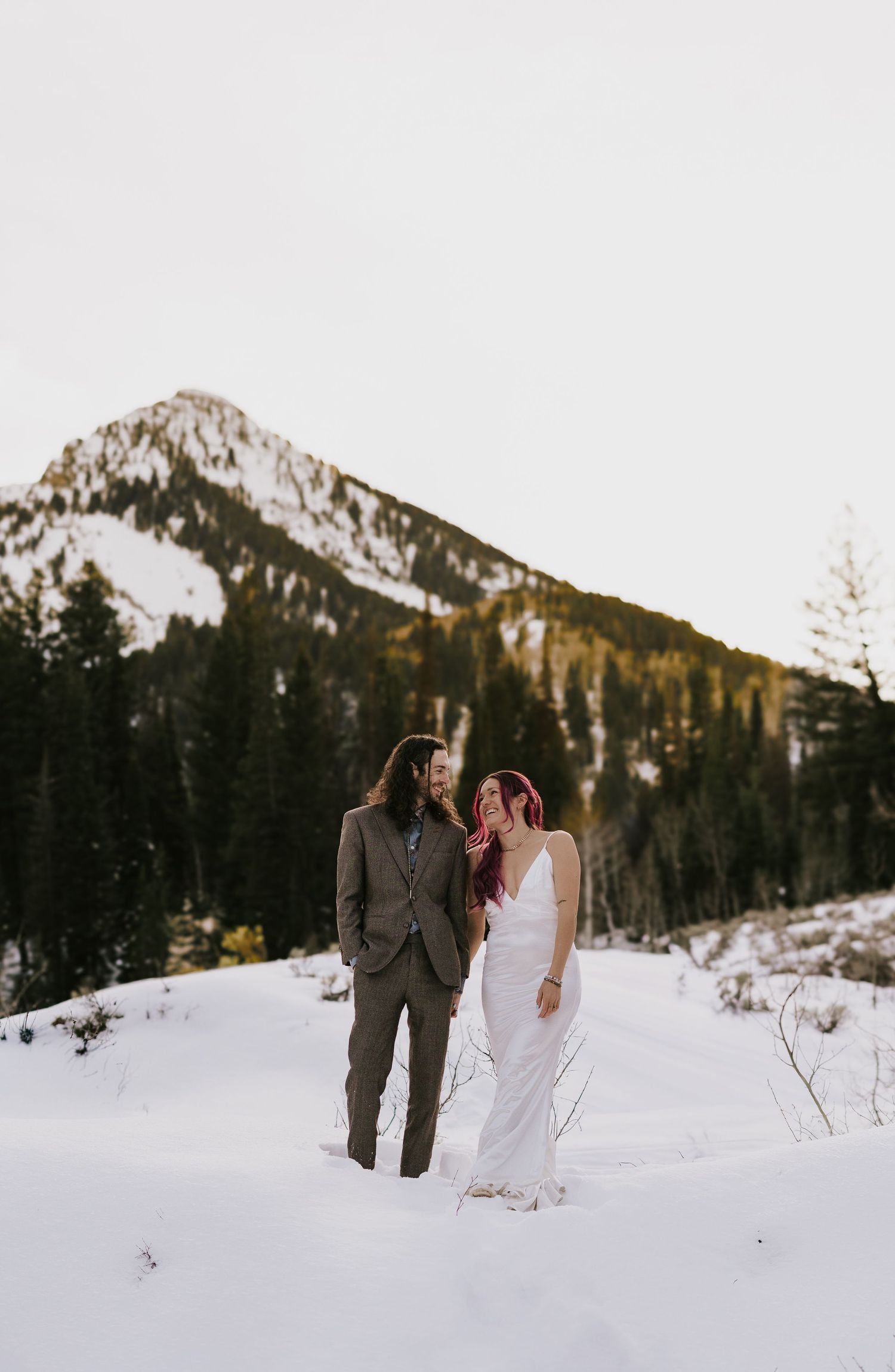 A couple in wedding attire stands together in a snowy mountain landscape with evergreen trees and peaks in the background.