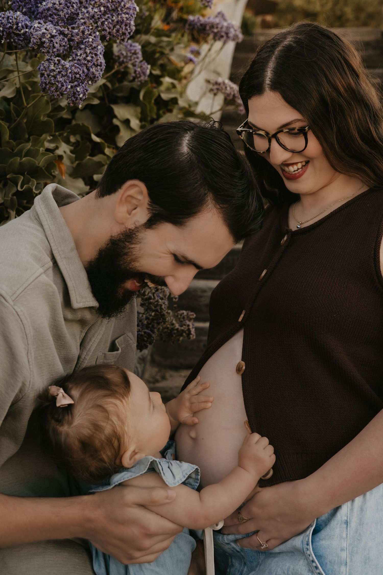 A couple smiles at their baby outdoors near purple flowers, with the woman showing a pregnant belly.