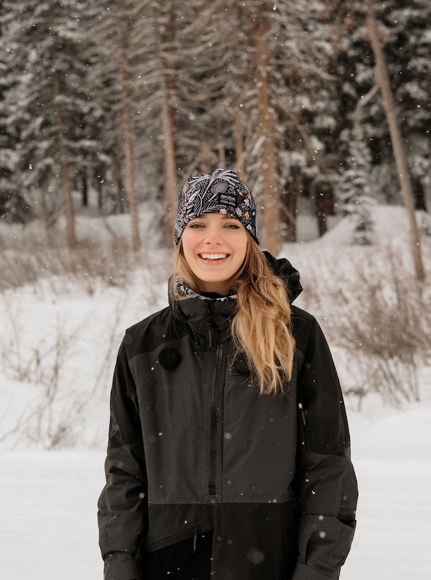 Person in a black winter coat and patterned beanie smiling while standing in a snowy forest setting.