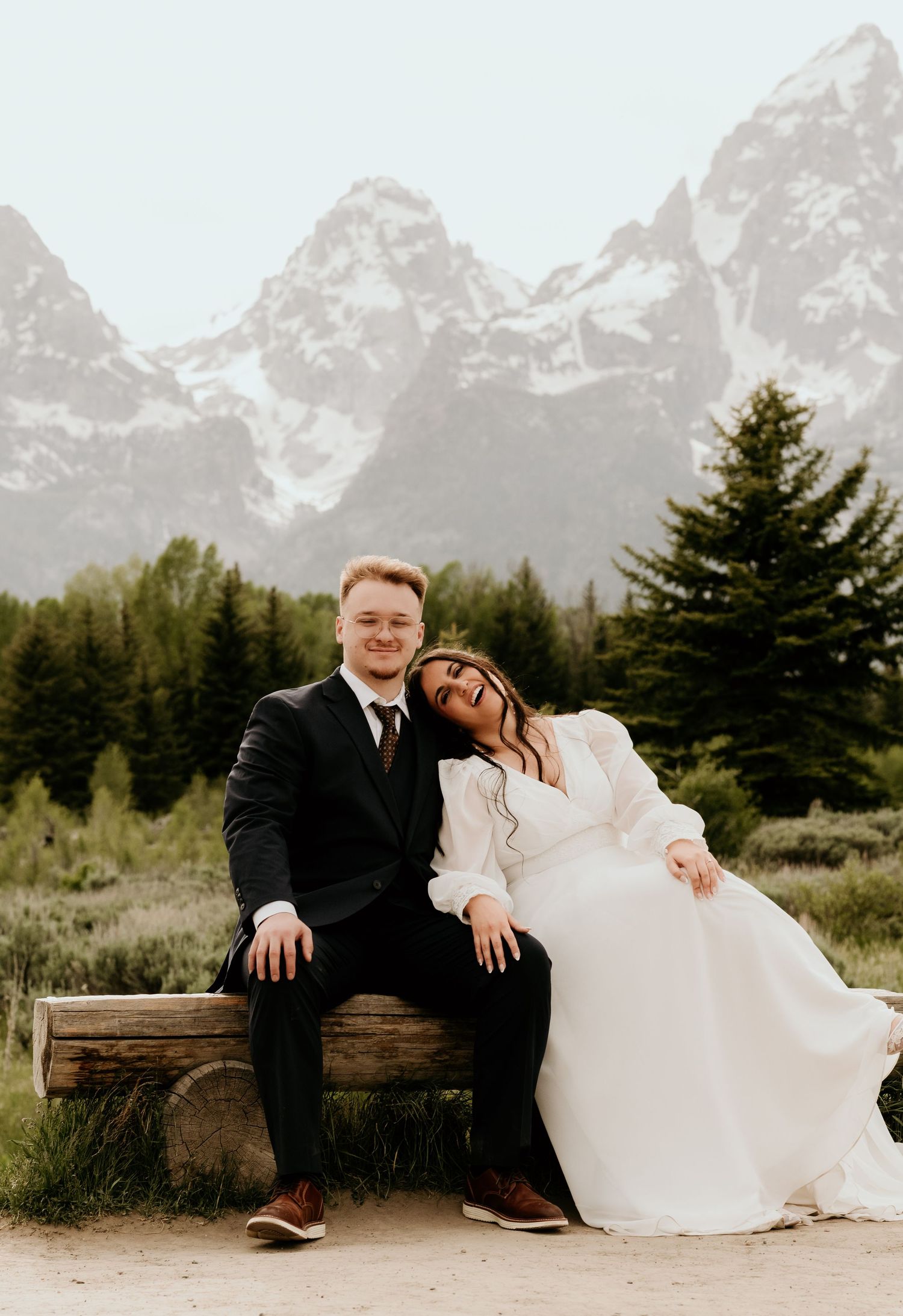 A couple in wedding attire sits on a log bench with the Grand Teton mountains and pine trees in the background.