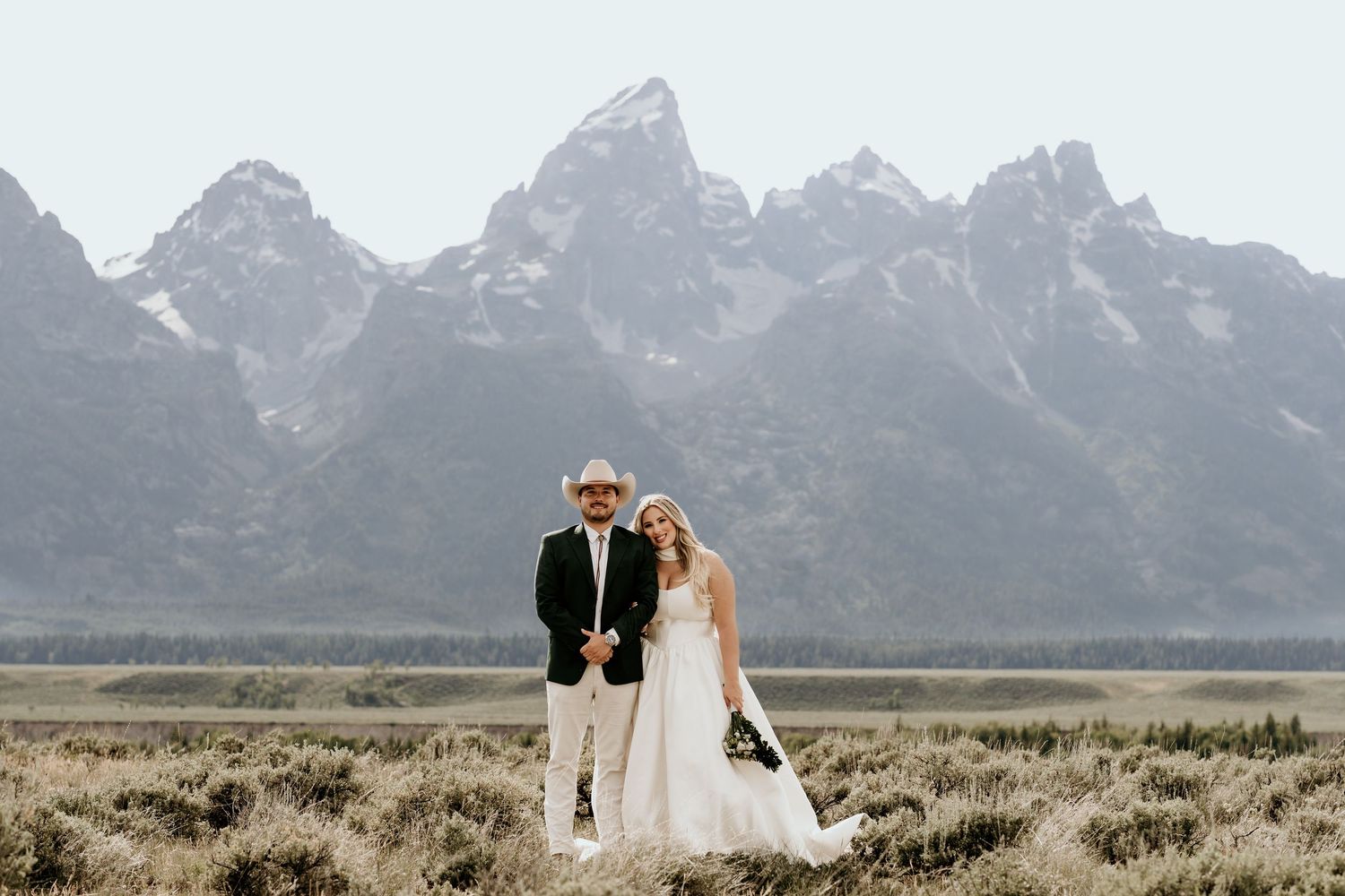 Bride and groom pose before the Grand Teton mountains in a vast Wyoming meadow on their wedding day.