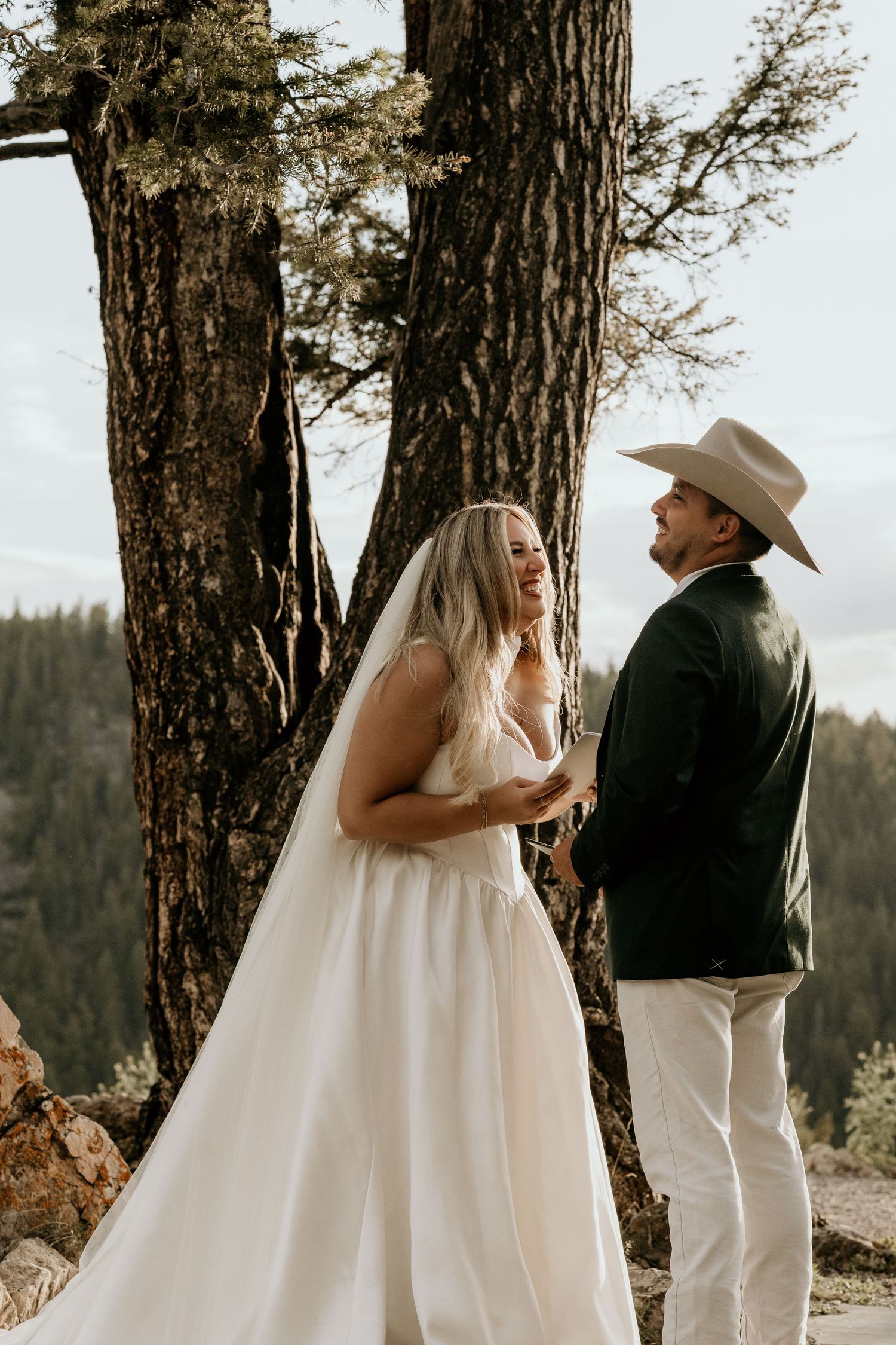 A bride in a white gown and veil exchanges vows with a groom in a cowboy hat outdoors among tall trees.