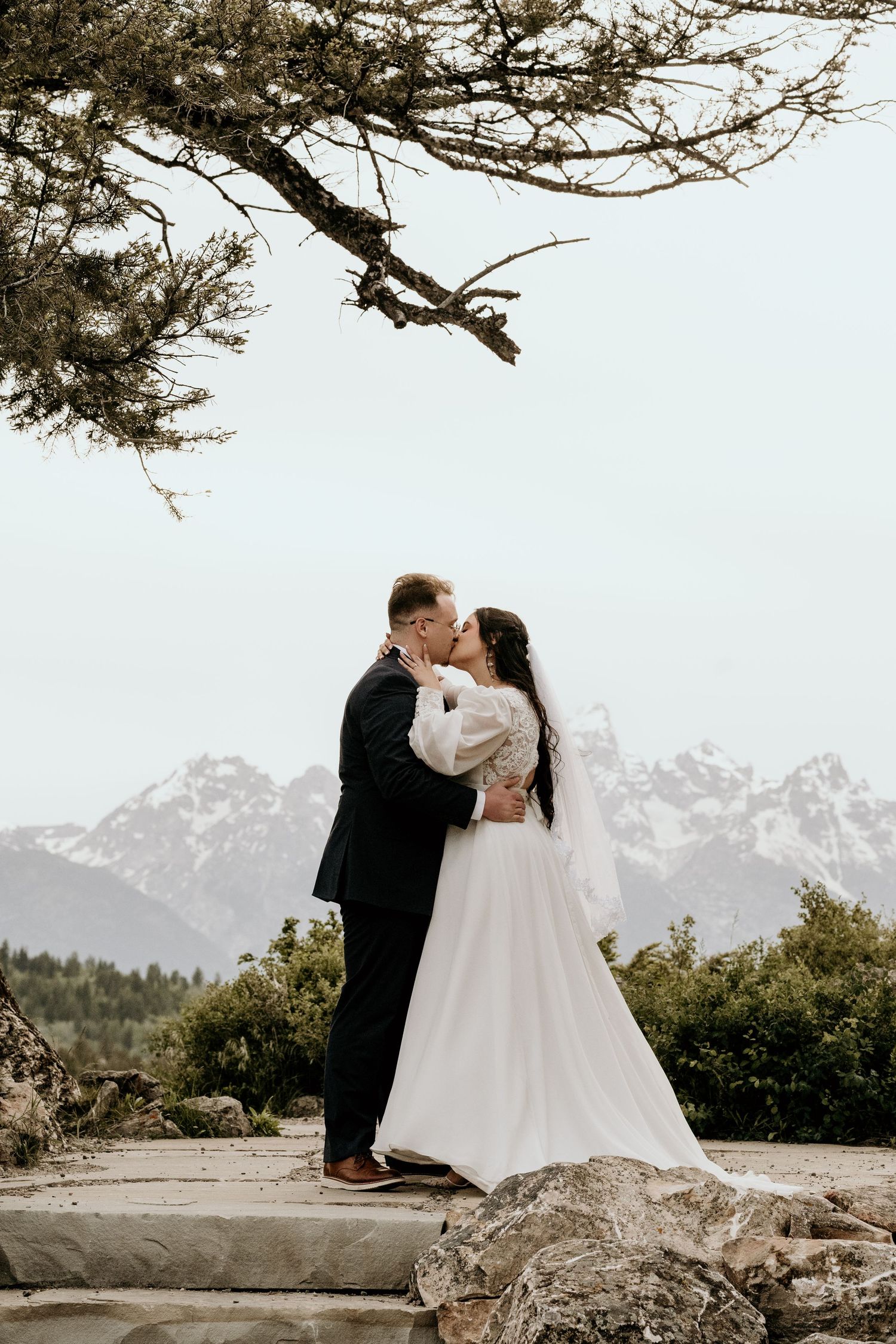 Bride and groom kissing with snow-capped Grand Teton mountains backdrop during outdoor wedding ceremony.