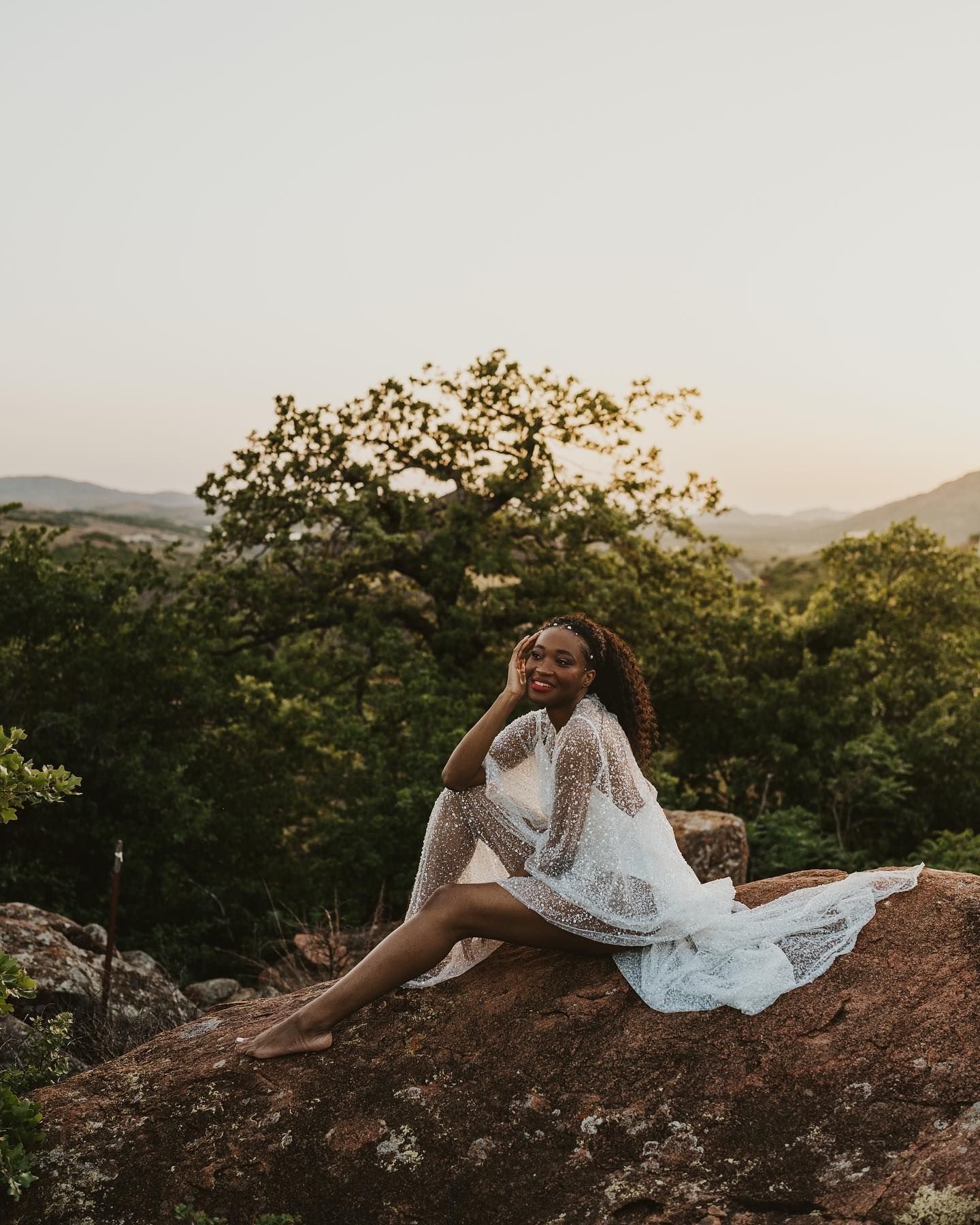 Bride in white dress reclines on rocky overlook during sunset.
