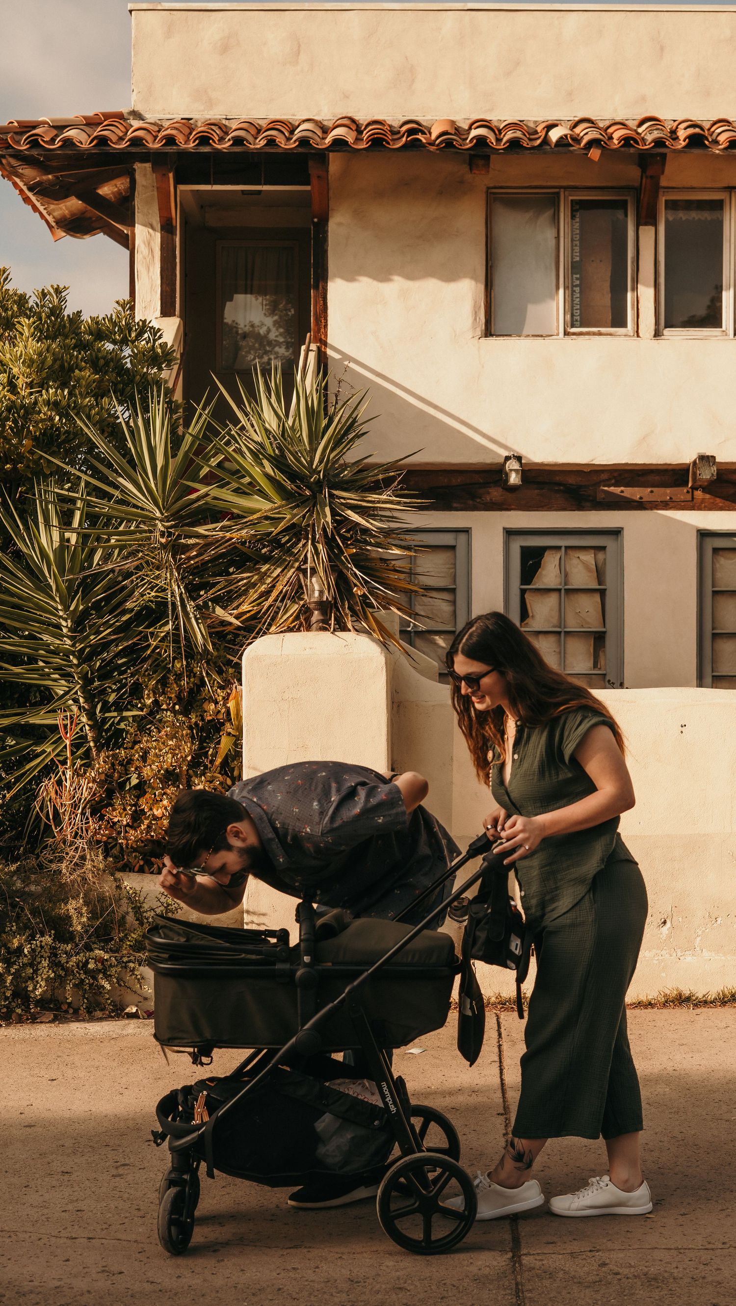 Parent adjusts stroller in front of stucco building during golden hour sunset.