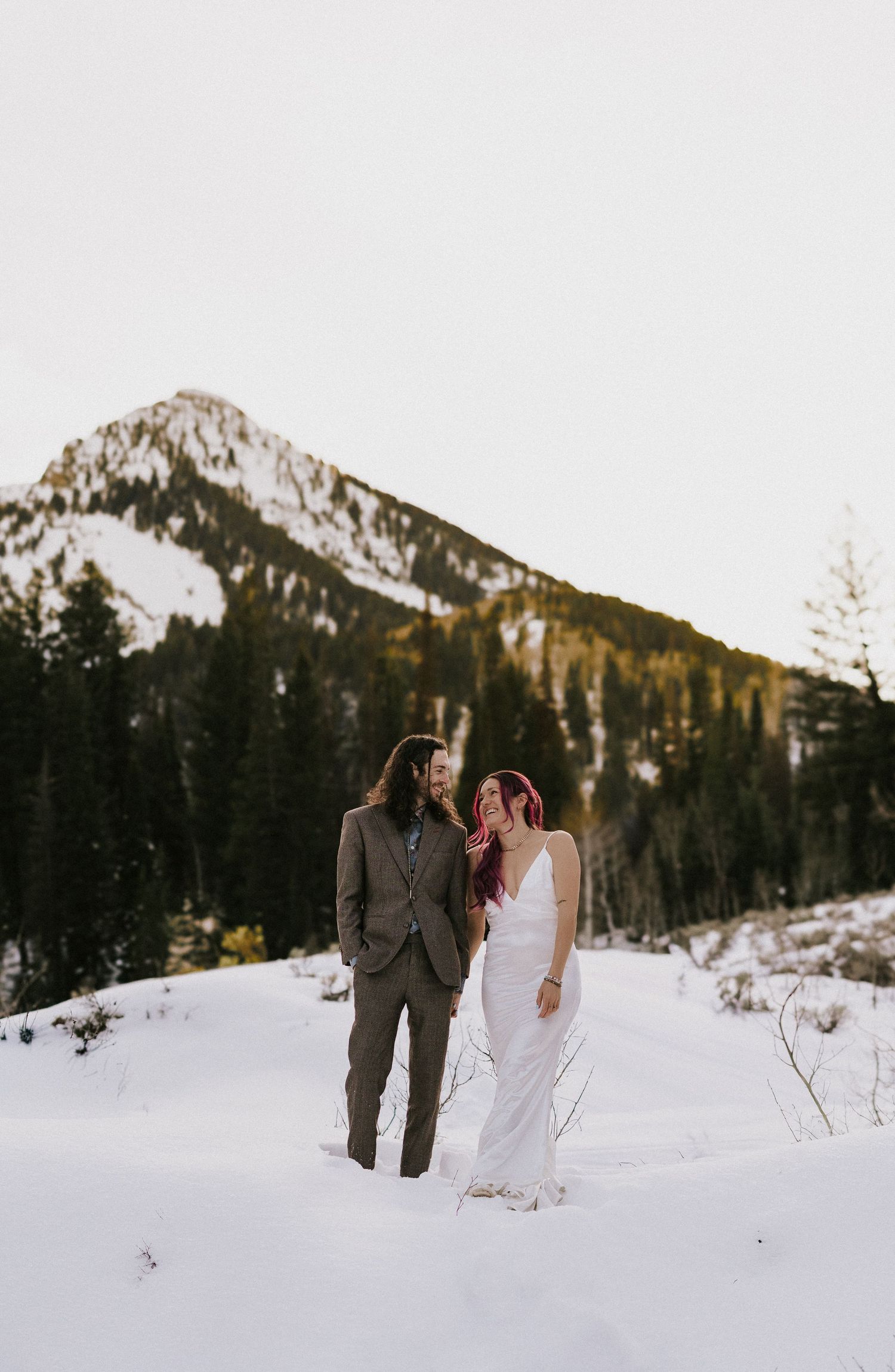 A couple walks through a snowy mountain landscape with evergreen trees and peaks in the background.
