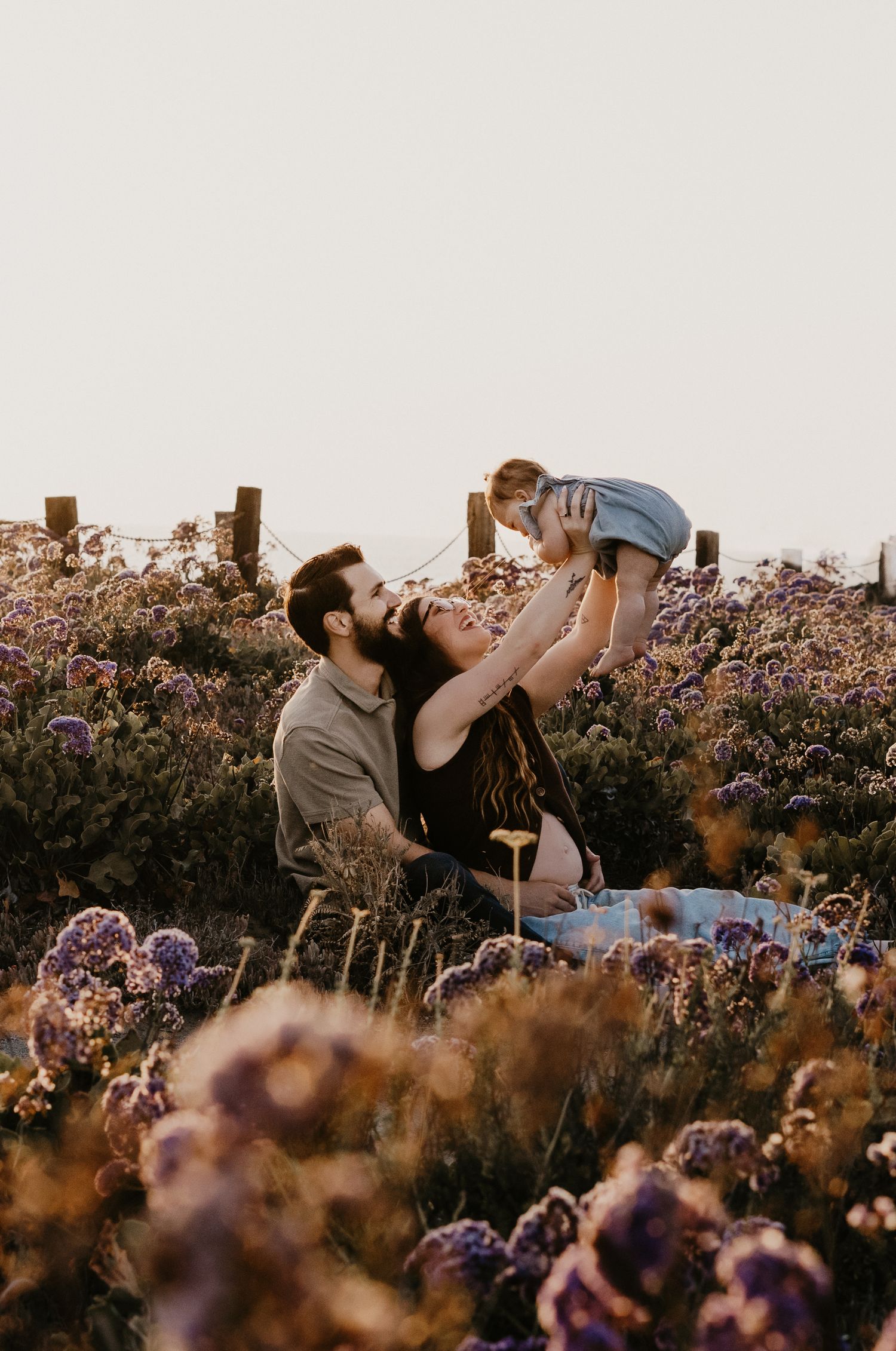A couple sits in a purple wildflower field, joyfully lifting their baby in golden hour light.