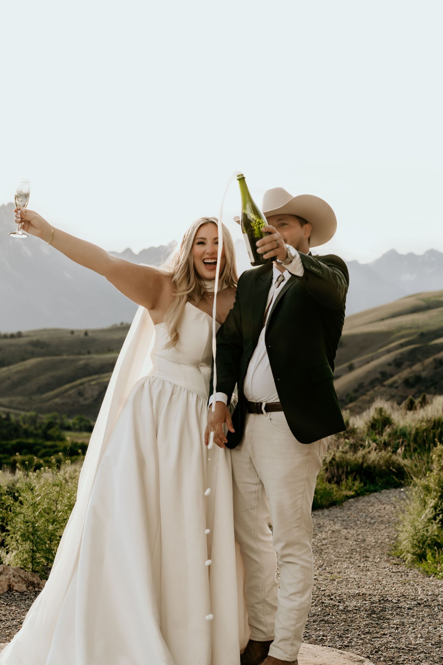 Joyful bride and groom celebrate with champagne toast on scenic mountain overlook, groom wearing cowboy hat.