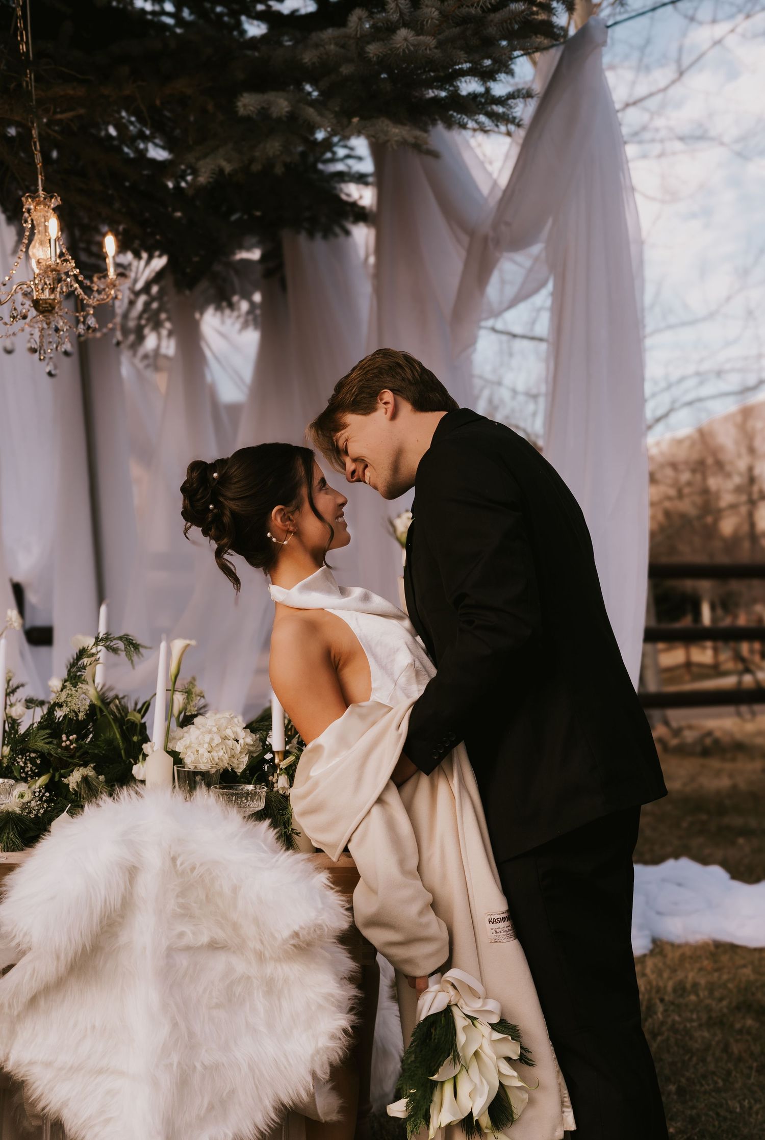 Romantic winter wedding couple in black and white attire sharing intimate moments outdoors with fur wrap and florals.