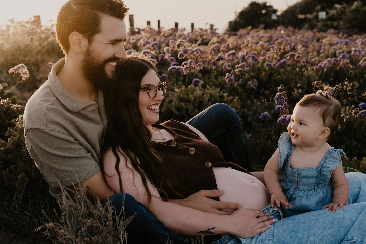 Happy family sitting in a flower field at golden hour, parents smiling at their baby girl in a blue dress.