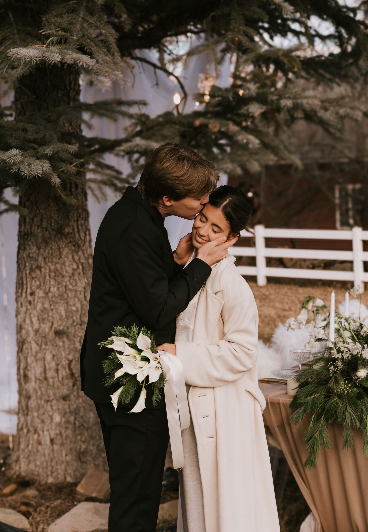 A romantic winter wedding moment with a couple embracing outdoors near a white fence and evergreen trees.
