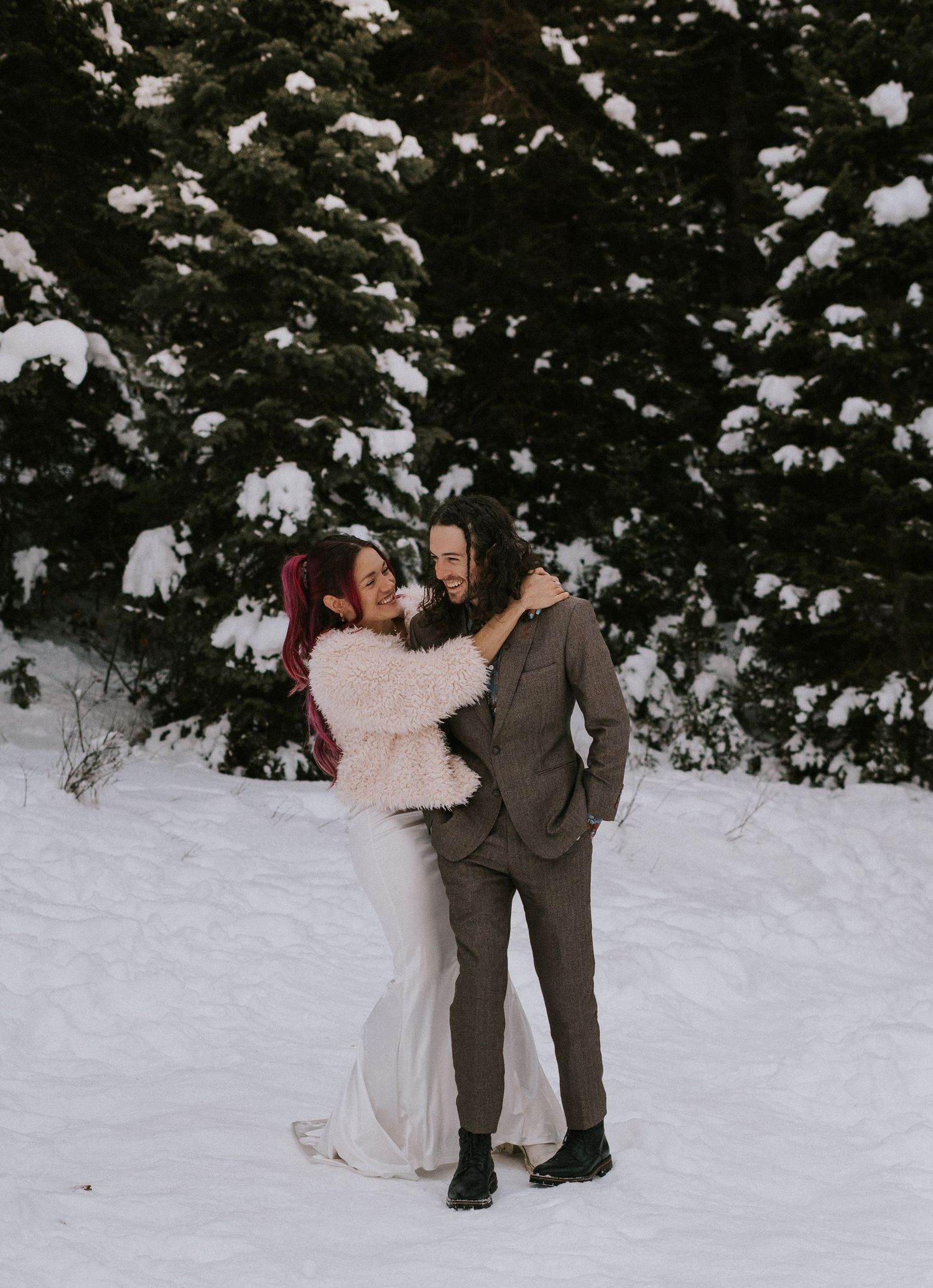 Couple plays in fresh snow surrounded by evergreen trees in winter.