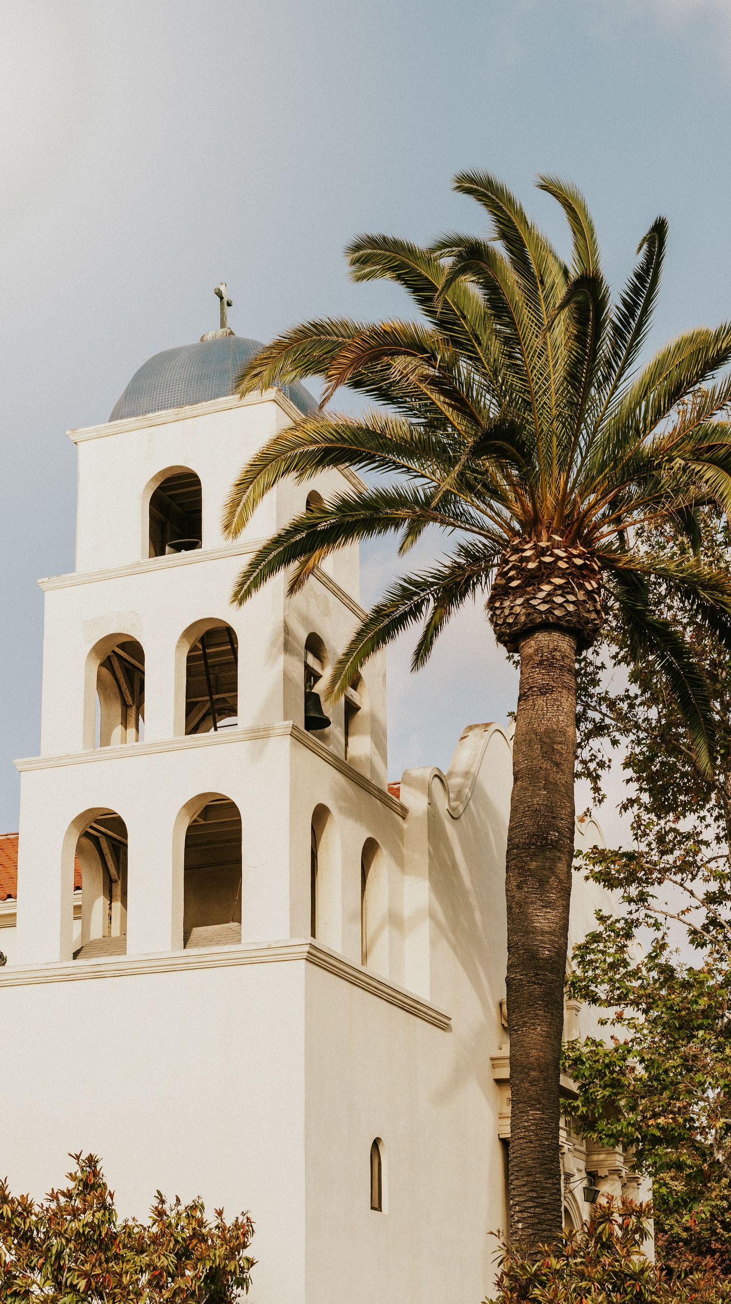 White Spanish mission-style bell tower with palm trees against blue sky.