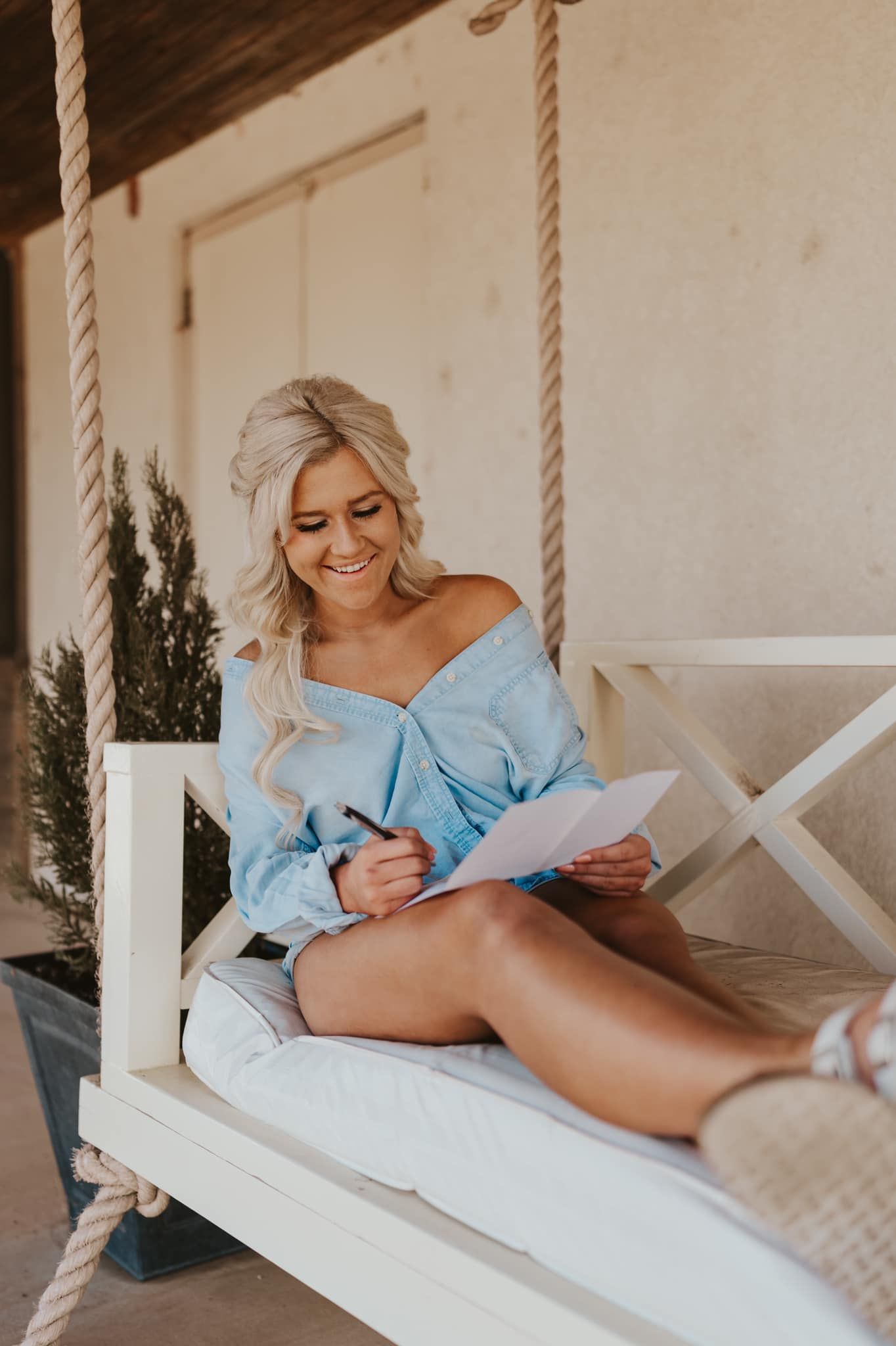 A person lounges comfortably on a hanging white porch swing while writing in natural light.