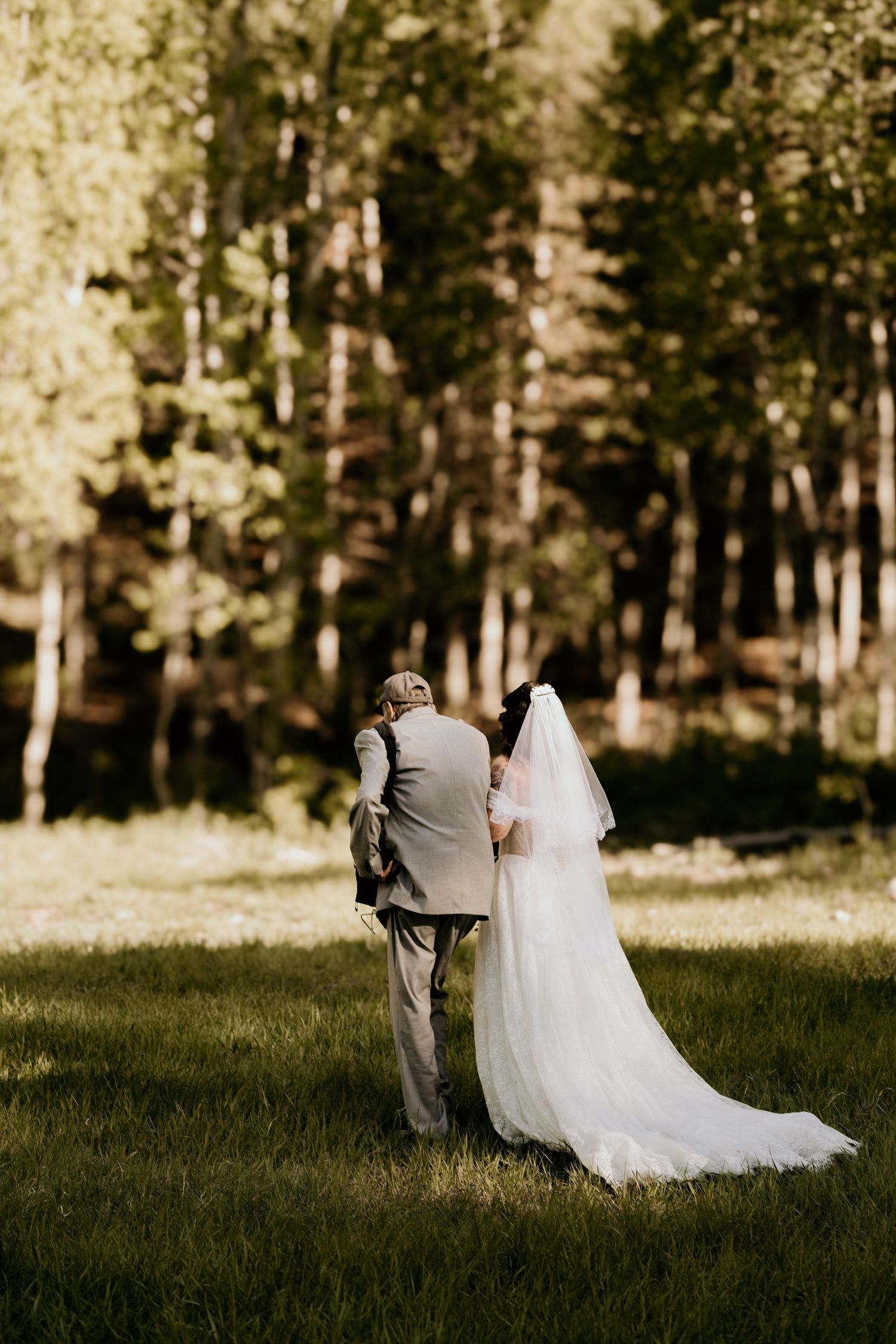 Bride in white gown and veil walks with escort through sunlit forest clearing during outdoor wedding ceremony.