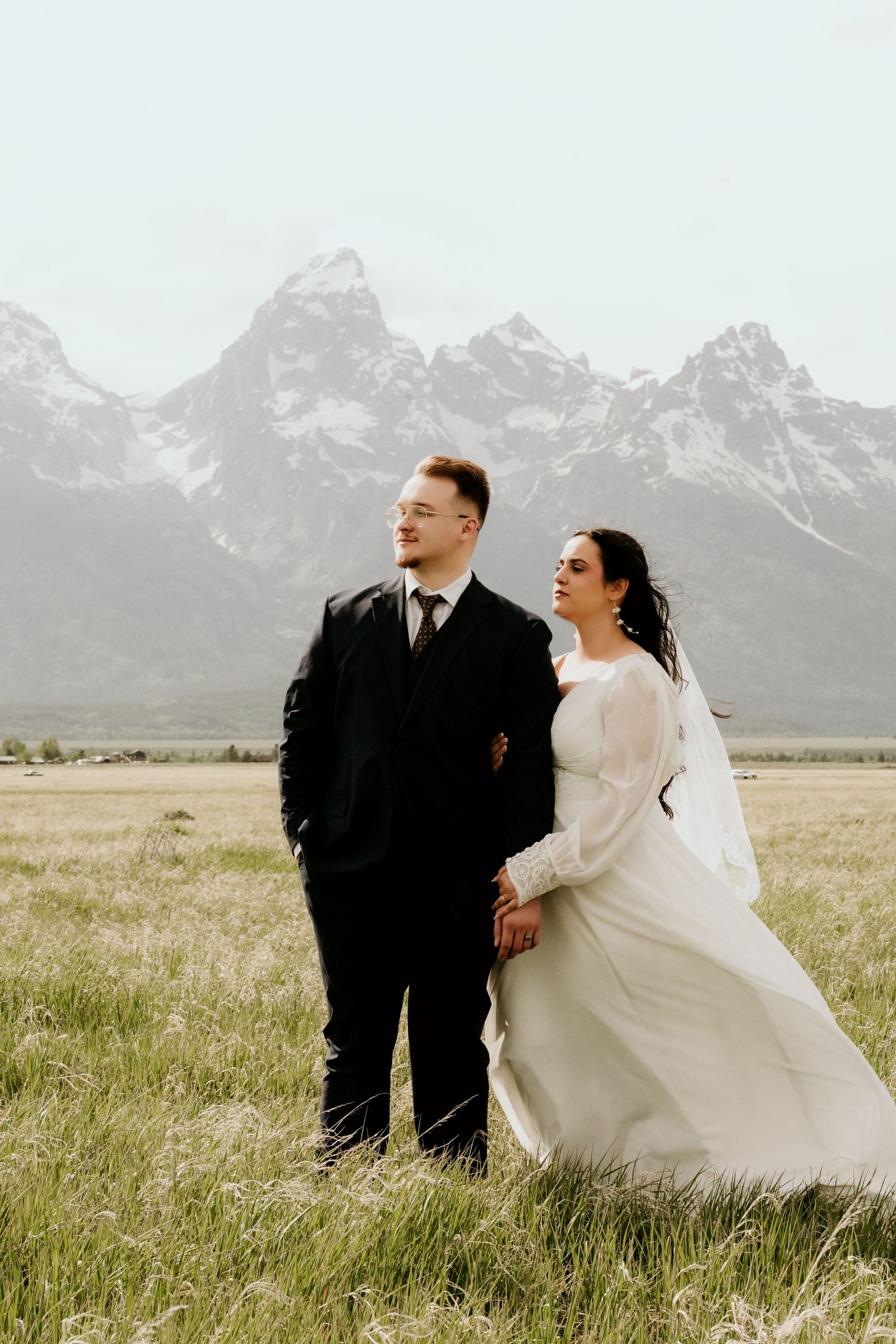 Bride and groom pose in a green meadow with snow-capped Grand Teton mountains in the background.