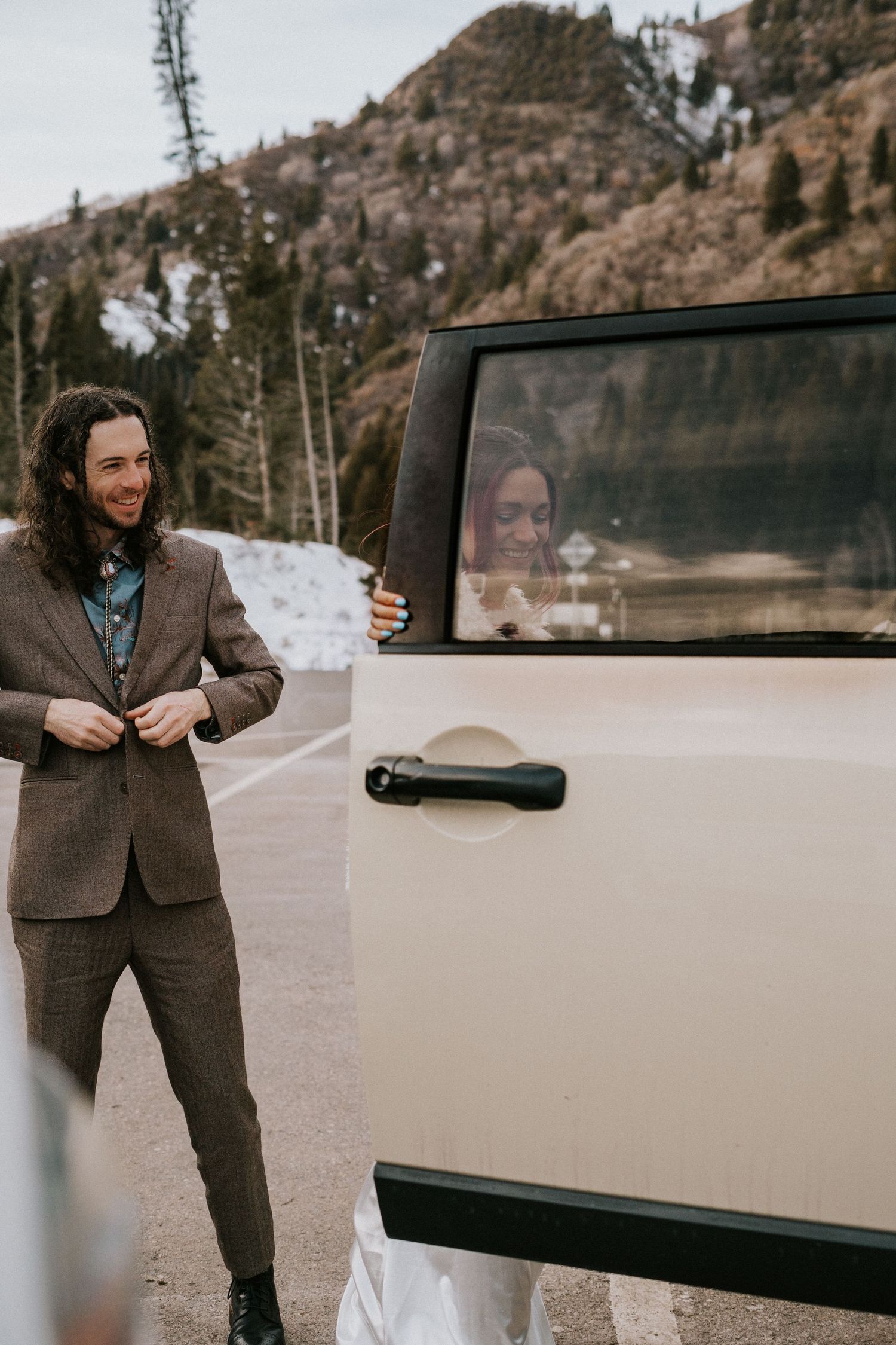 Person in brown suit standing next to vintage car in snowy mountain setting during winter photoshoot.