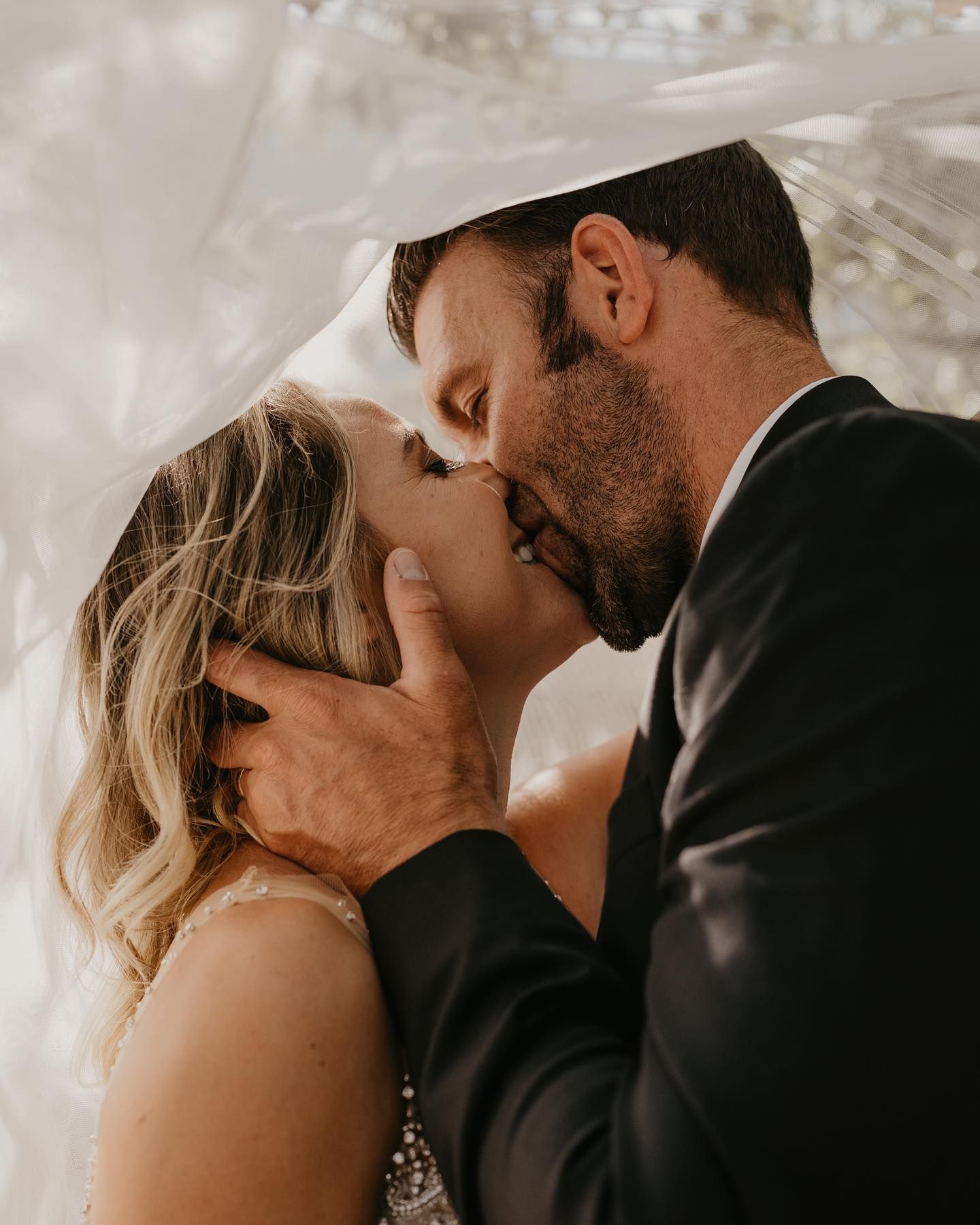 Romantic wedding couple sharing an intimate kiss under flowing veil during golden hour.