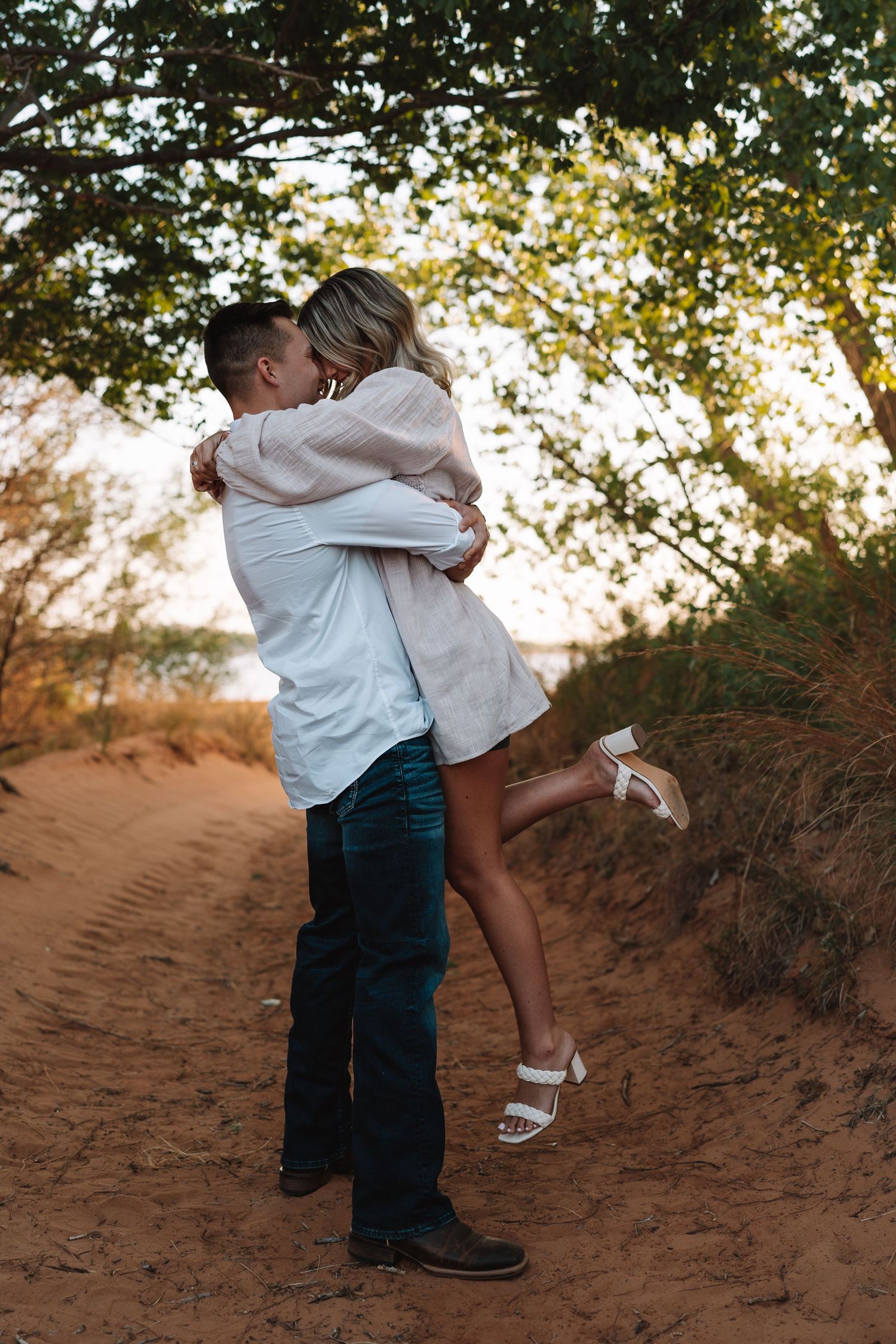 Playful couple embracing on sandy terrain with trees in the background during sunset.