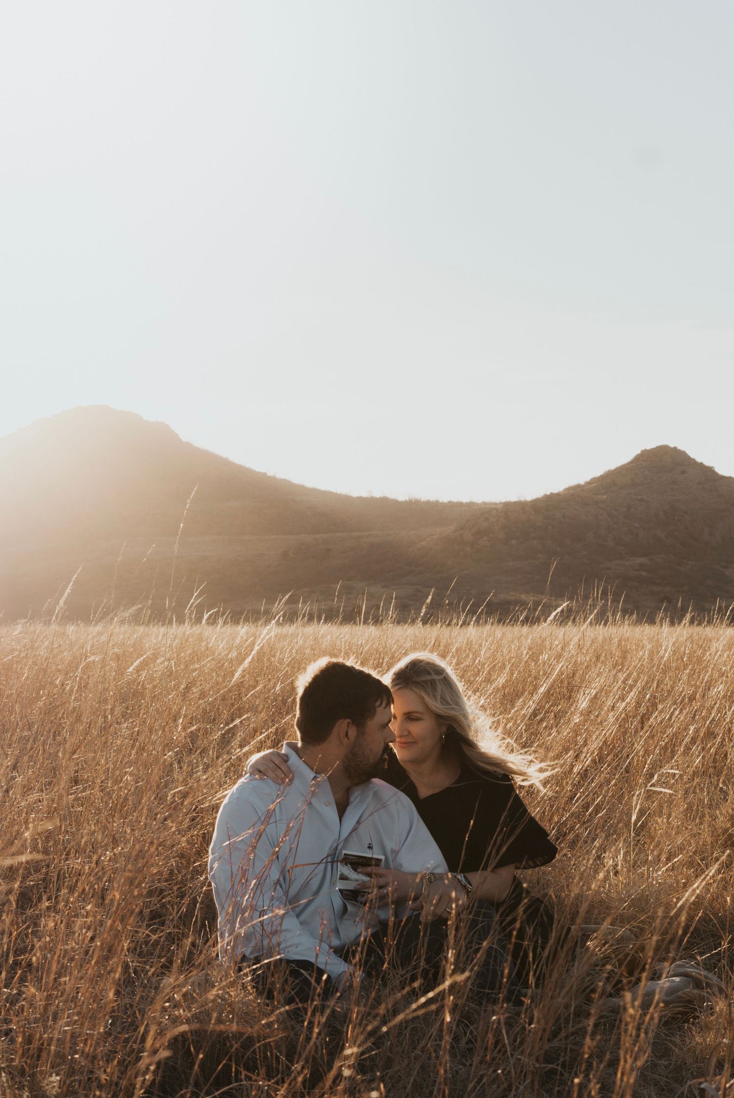 Couple in a golden wheat field at sunset with mountains in the background.