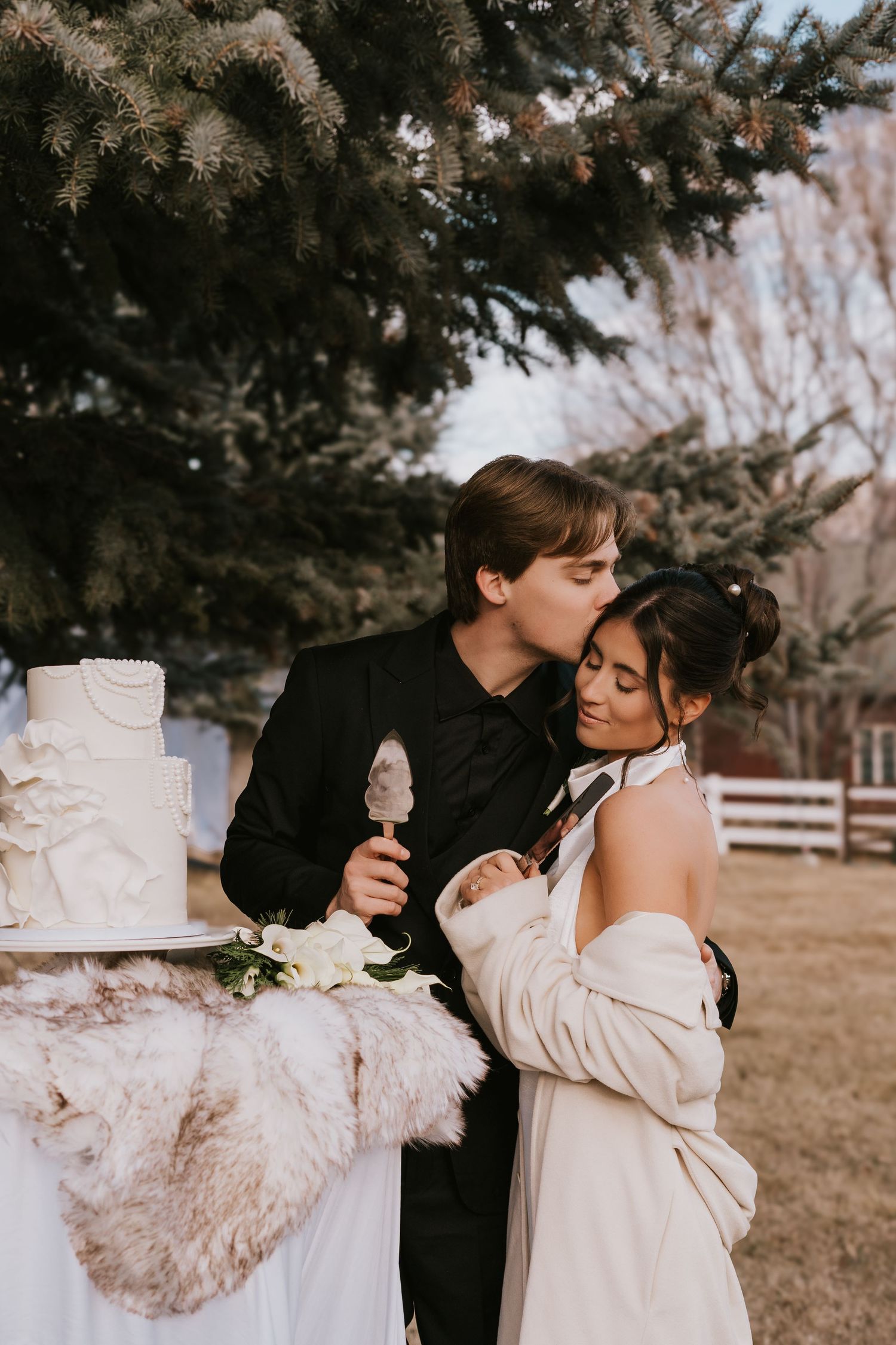 A romantic wedding moment outdoors with a white cake on display as a couple shares an intimate embrace near evergreen trees.