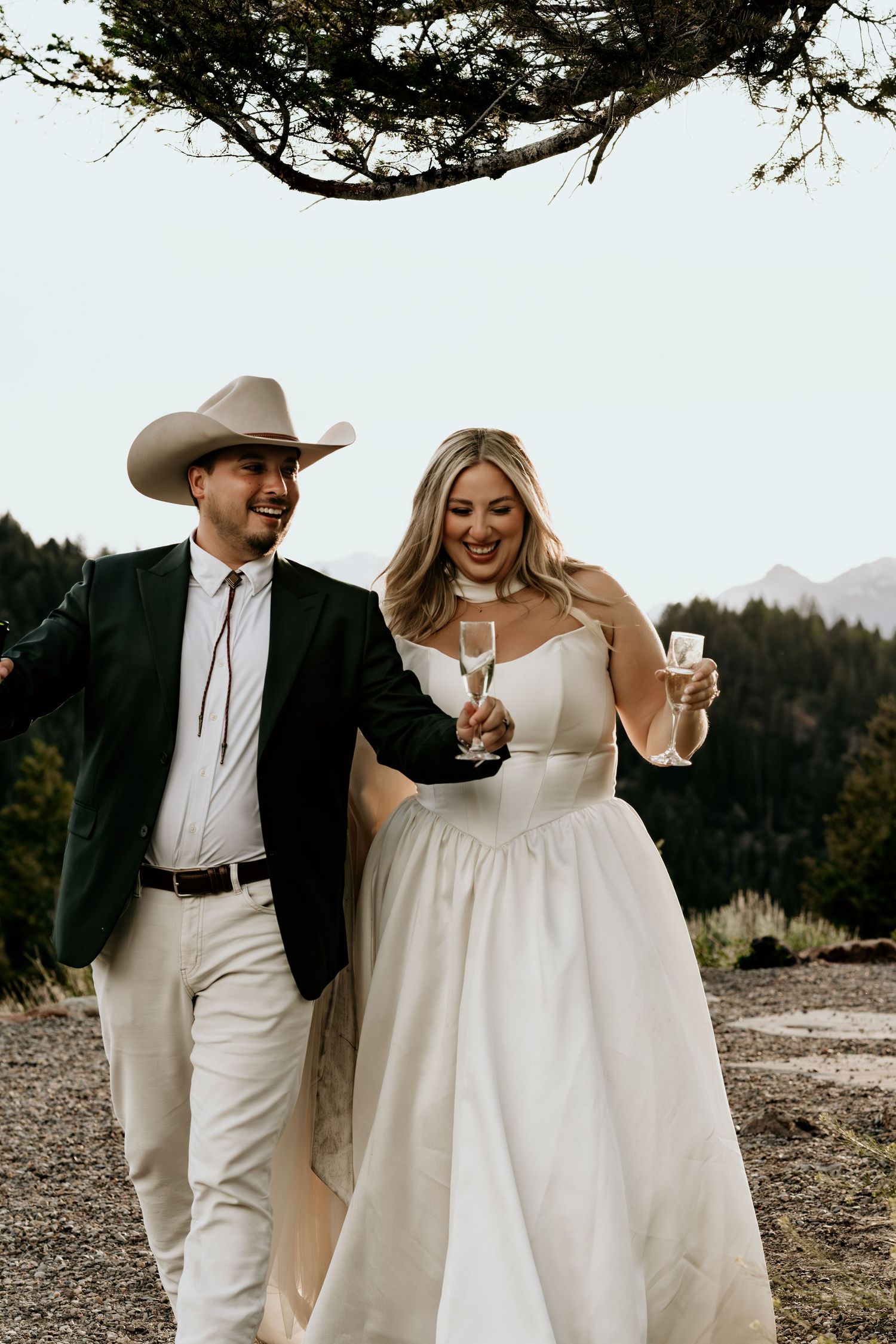 Joyful bride in white gown and groom in cowboy hat celebrate with champagne at an outdoor mountain wedding.