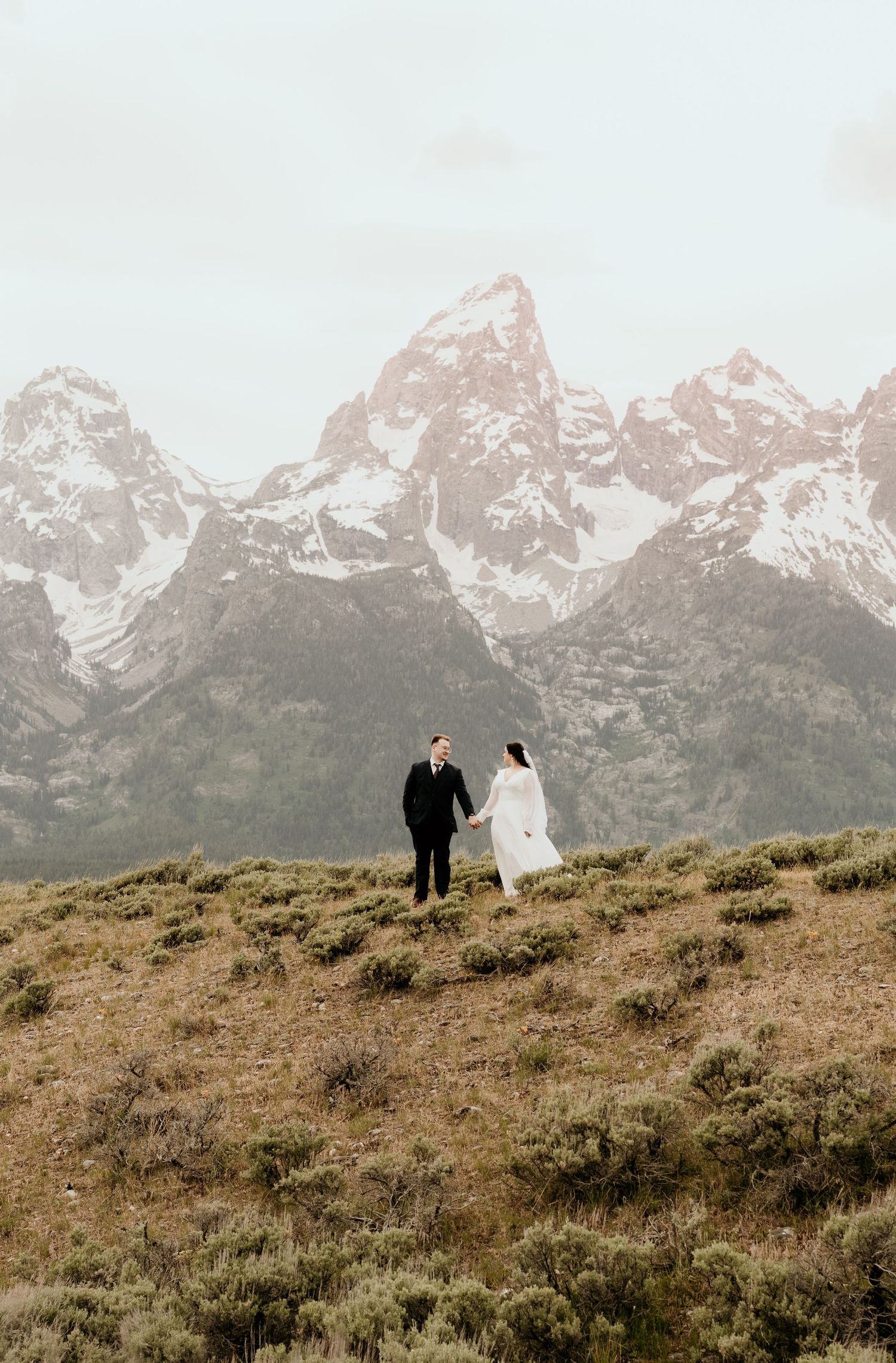 Couple holding hands in wedding attire with snow-capped Grand Teton mountains in the background.