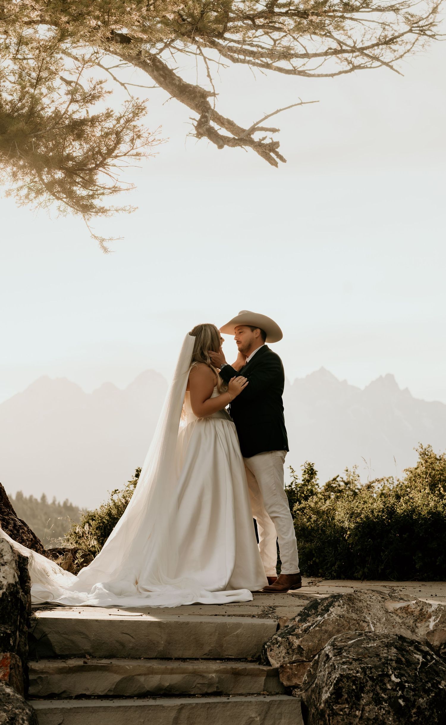Bride and groom share a romantic moment on stone steps with misty mountain views and a flowing white veil.