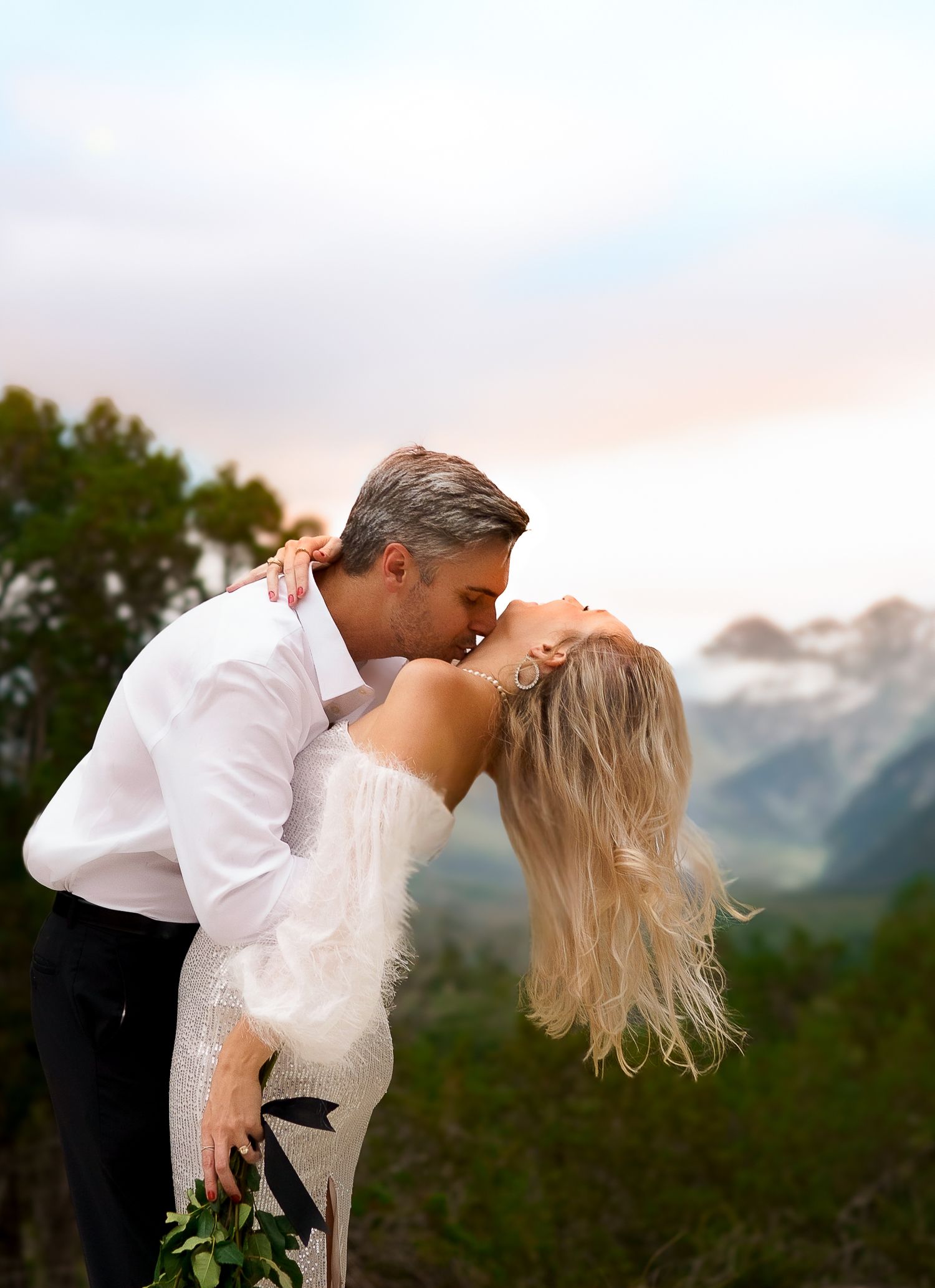 A couple shares a romantic kiss against a backdrop of majestic mountain peaks during sunset.