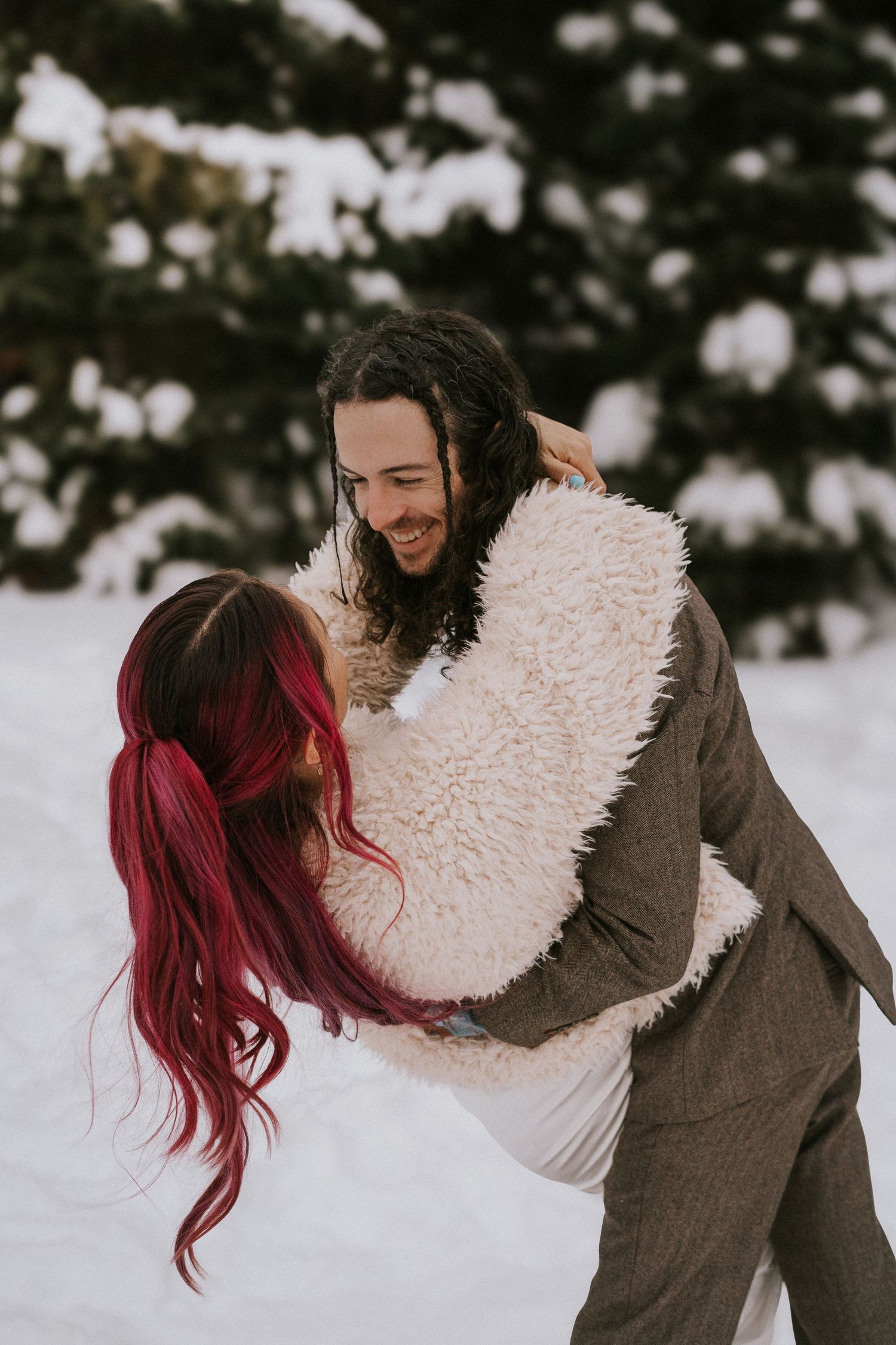 Playful couple in winter attire hugging in the snow with evergreen trees in background.