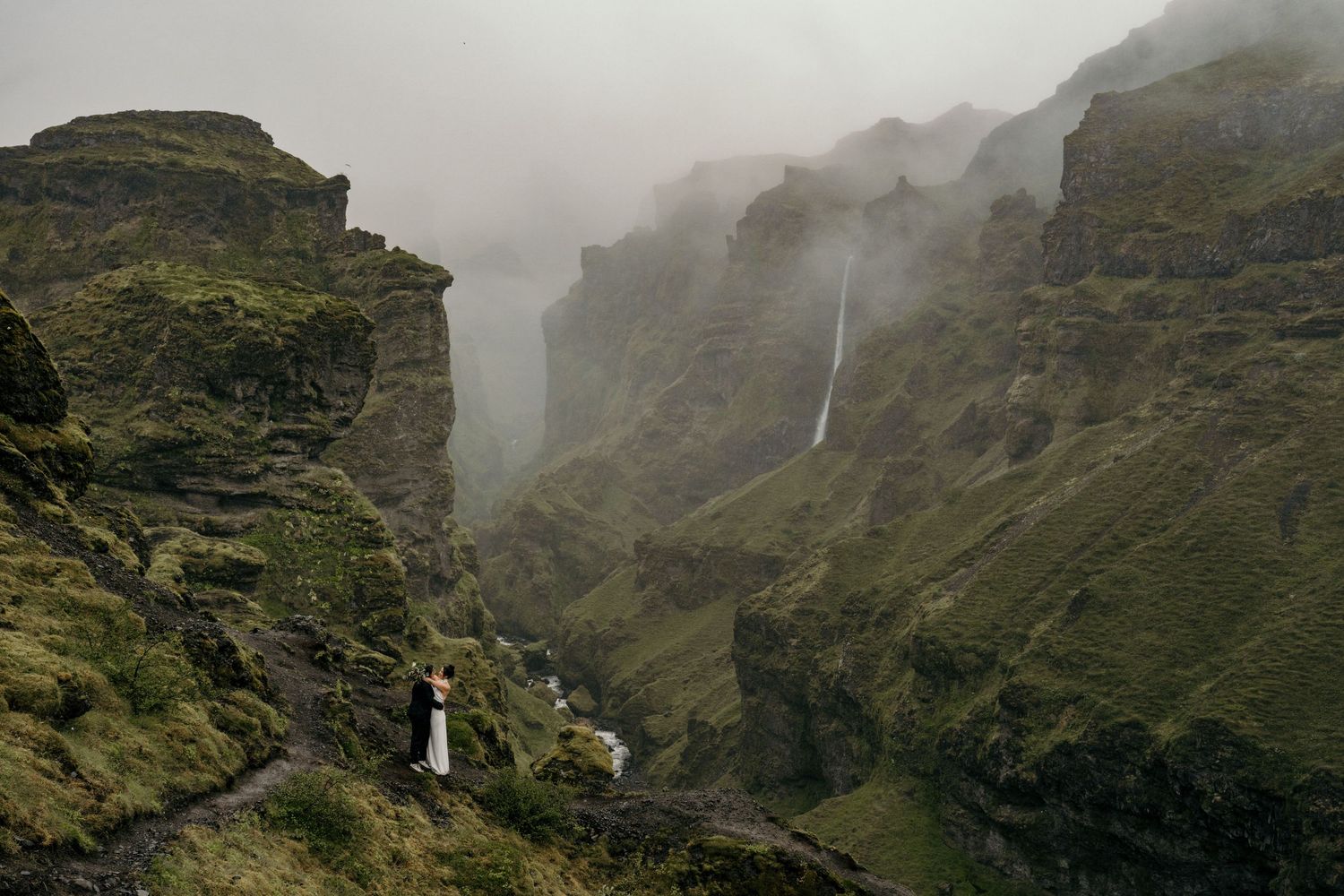 Couple eloping in a dramatic misty green canyon with a waterfall cascading down rugged cliffs in Iceland.