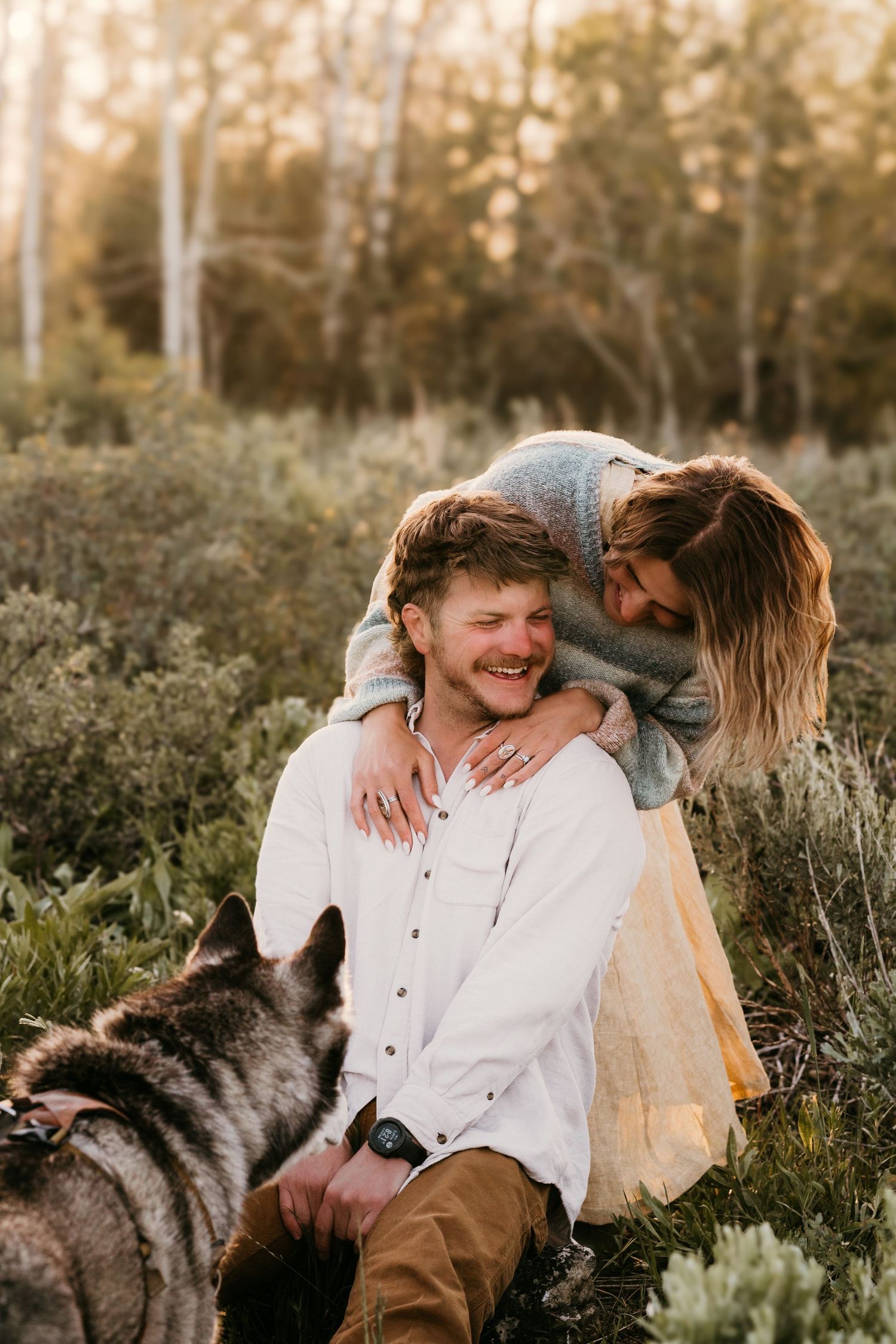 Couple sitting in a field at golden hour, woman hugging man from behind while a husky dog looks on.