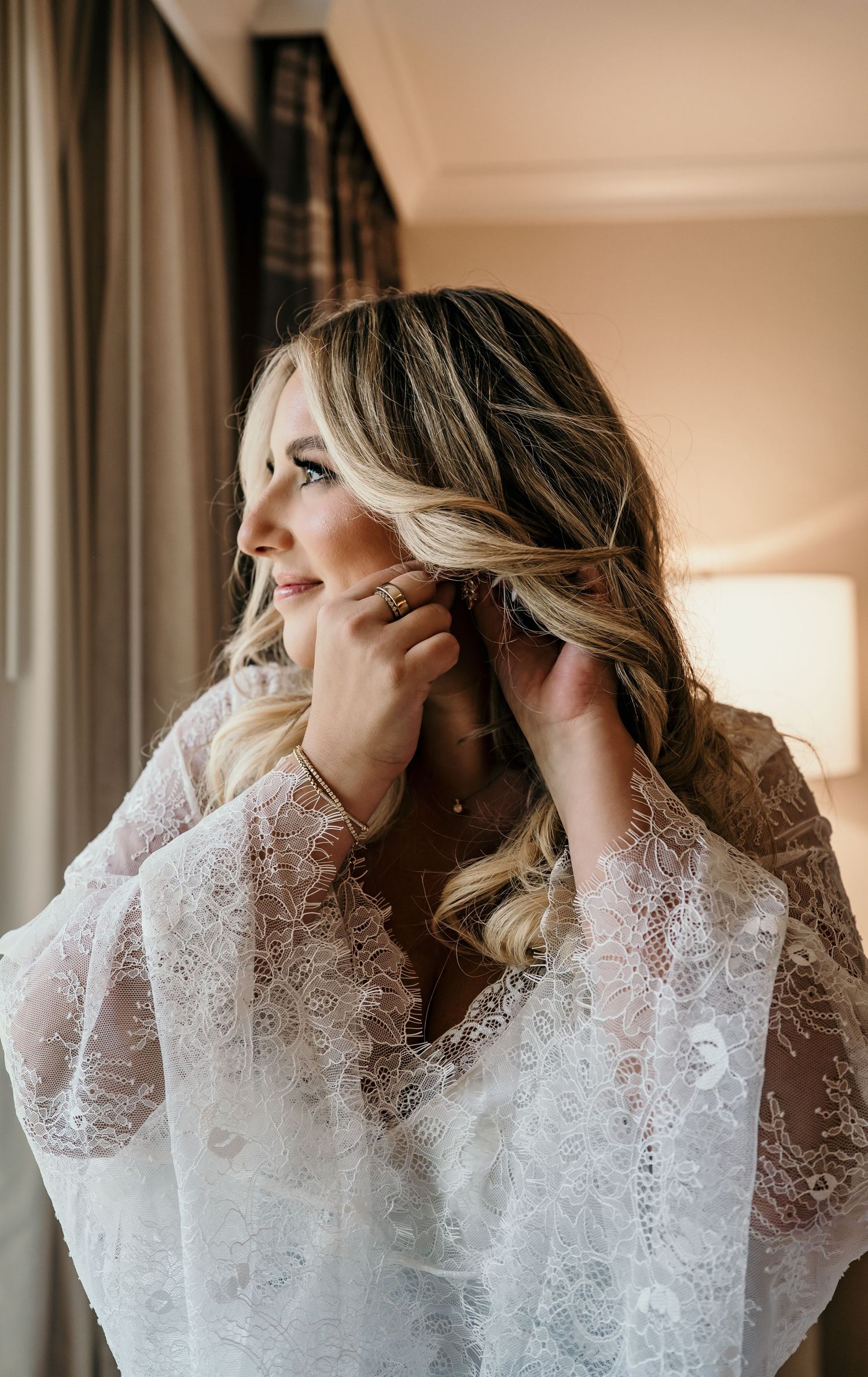 Bride in lace robe putting on earrings while getting ready, with soft natural light and wavy blonde hair.