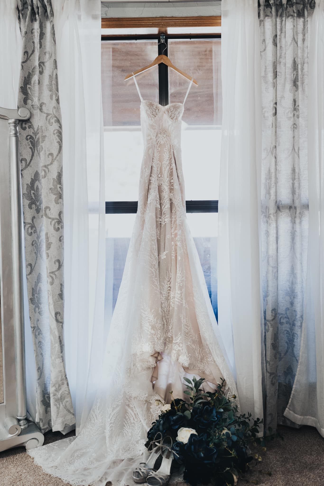 Elegant lace wedding dress hangs in window with floral bouquet at base and flowing white curtains framing the scene.