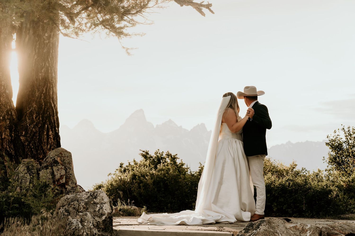 Bride and groom share a romantic moment at mountain overlook with misty Teton peaks at sunset.
