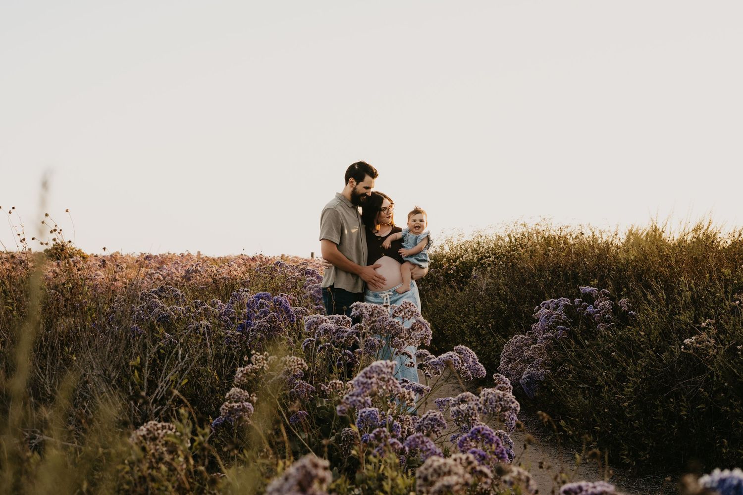 Family of three standing among purple wildflowers in a golden field at sunset.
