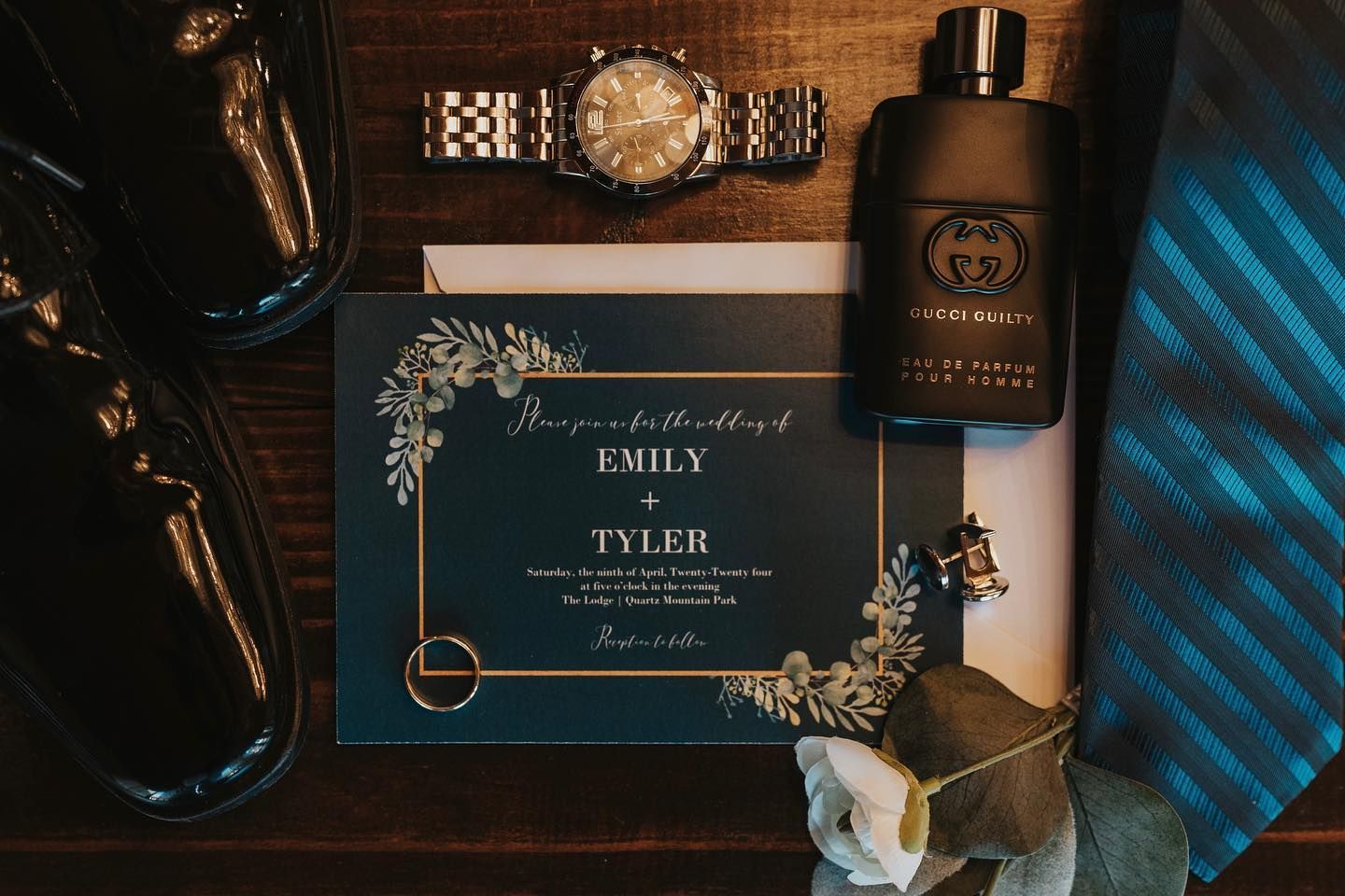 Elegant wedding details featuring blue and gold decor with invitations, rings, and floral arrangements on a wooden surface.
