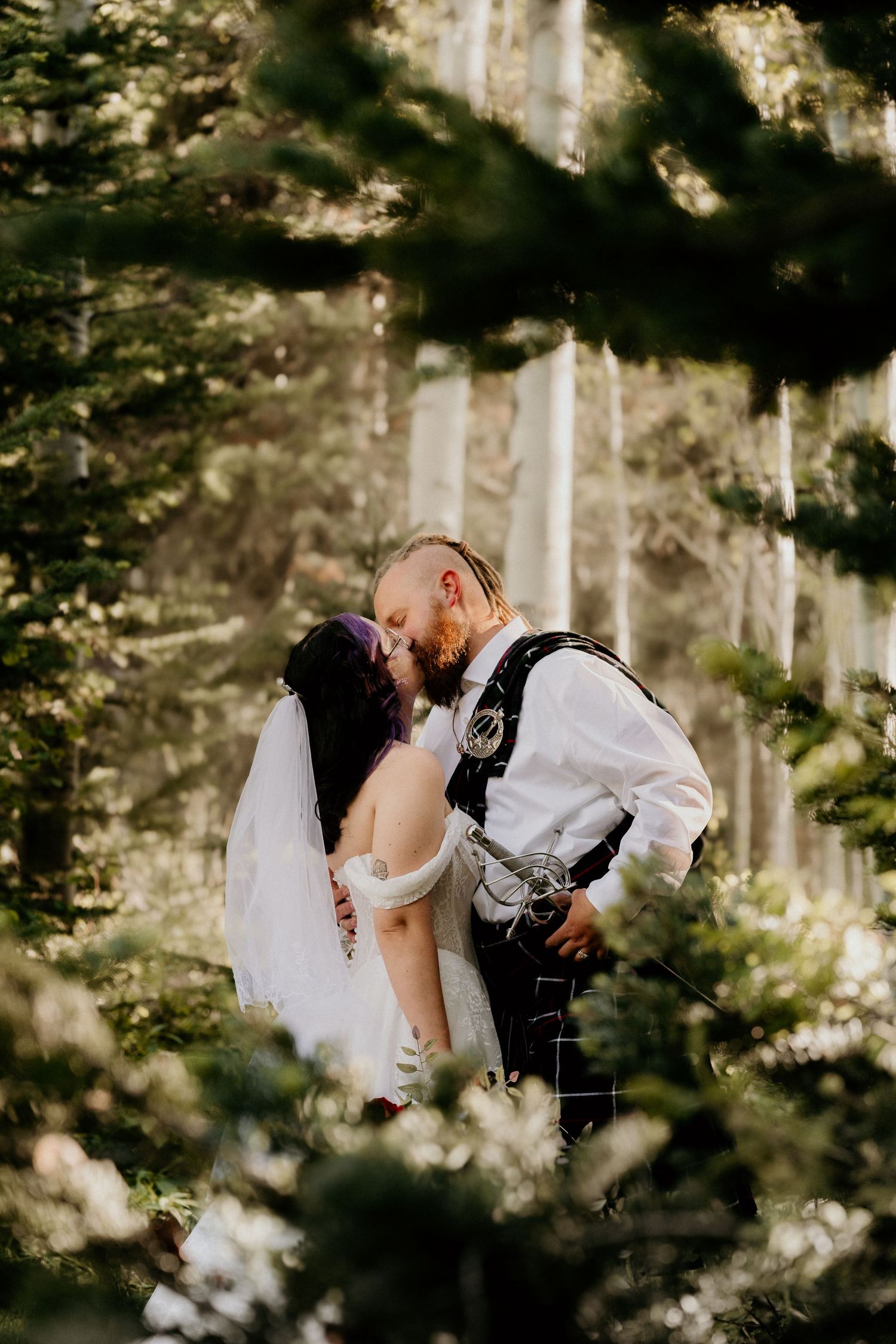A bride and groom share a romantic kiss surrounded by lush green pine trees in a forest wedding setting.