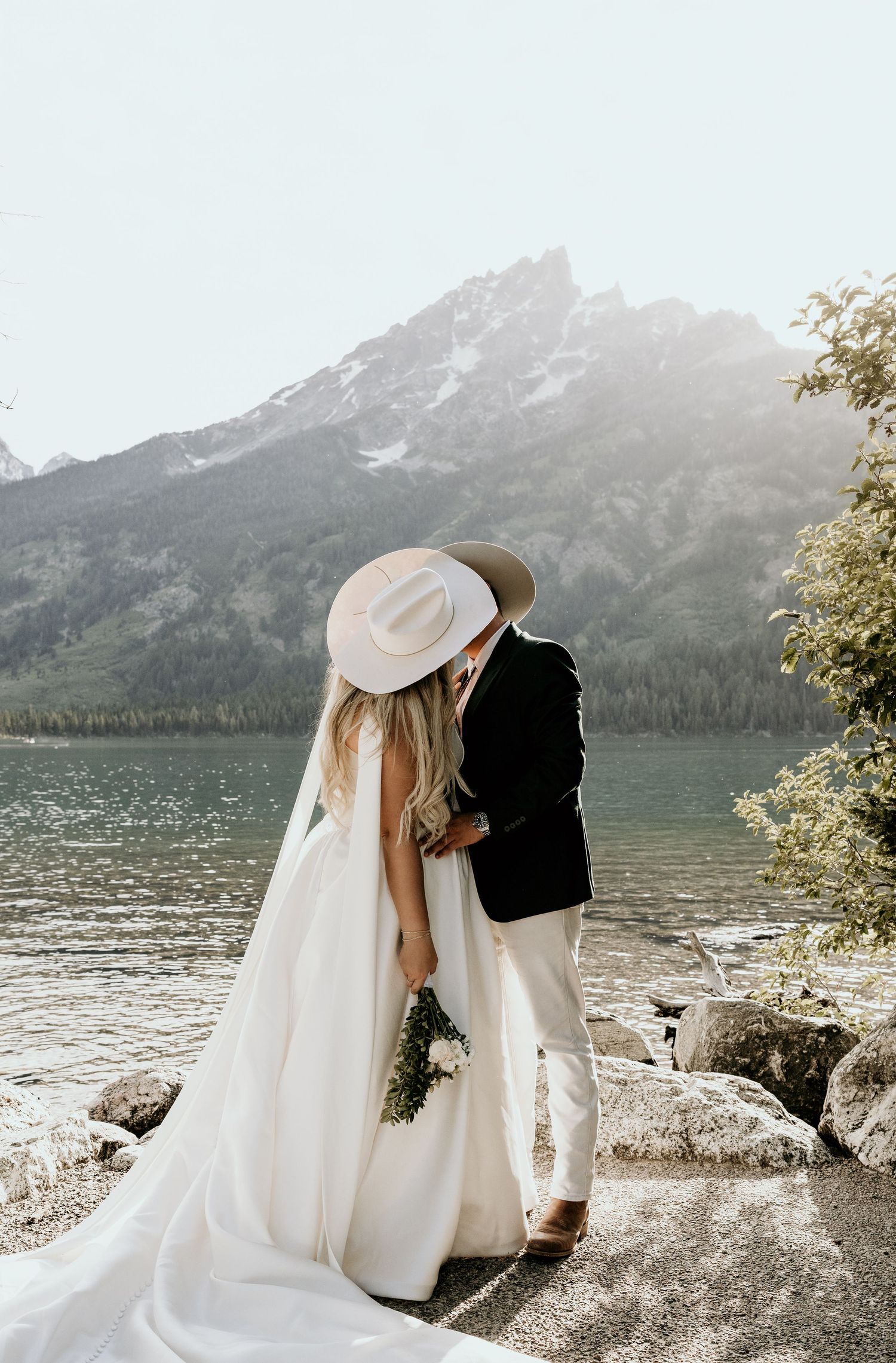 Bride and groom share a kiss by a mountain lake, with stunning peaks reflected in the water behind them.