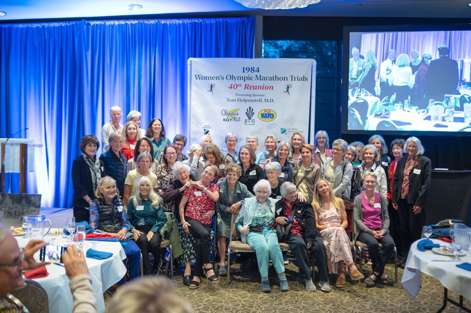 Group photo of women at the 1984 Women's Olympic Marathon Trials 40th reunion event in a banquet hall.