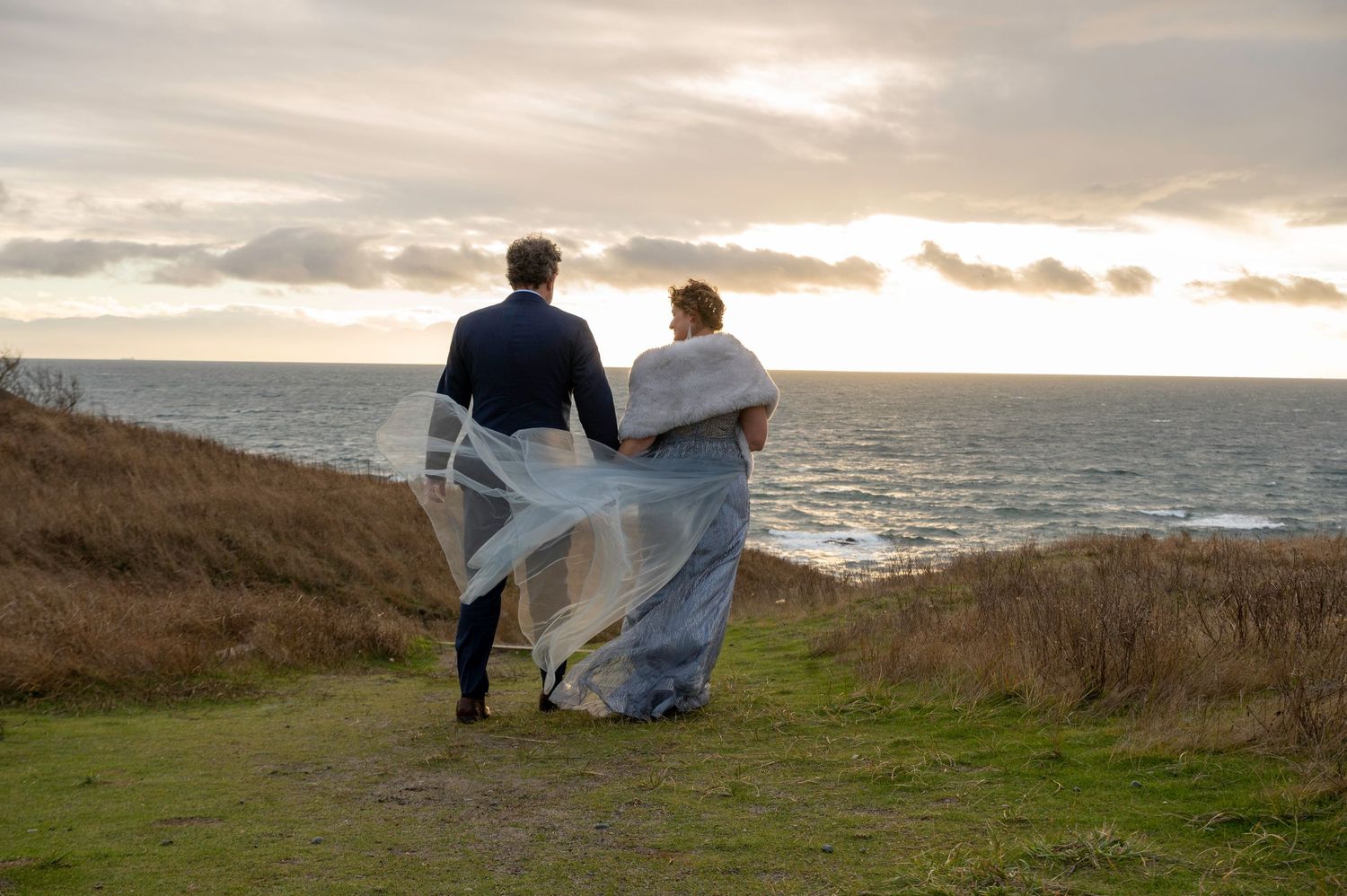 A couple in wedding attire stands together overlooking a dramatic coastal sunset on a grassy cliff.