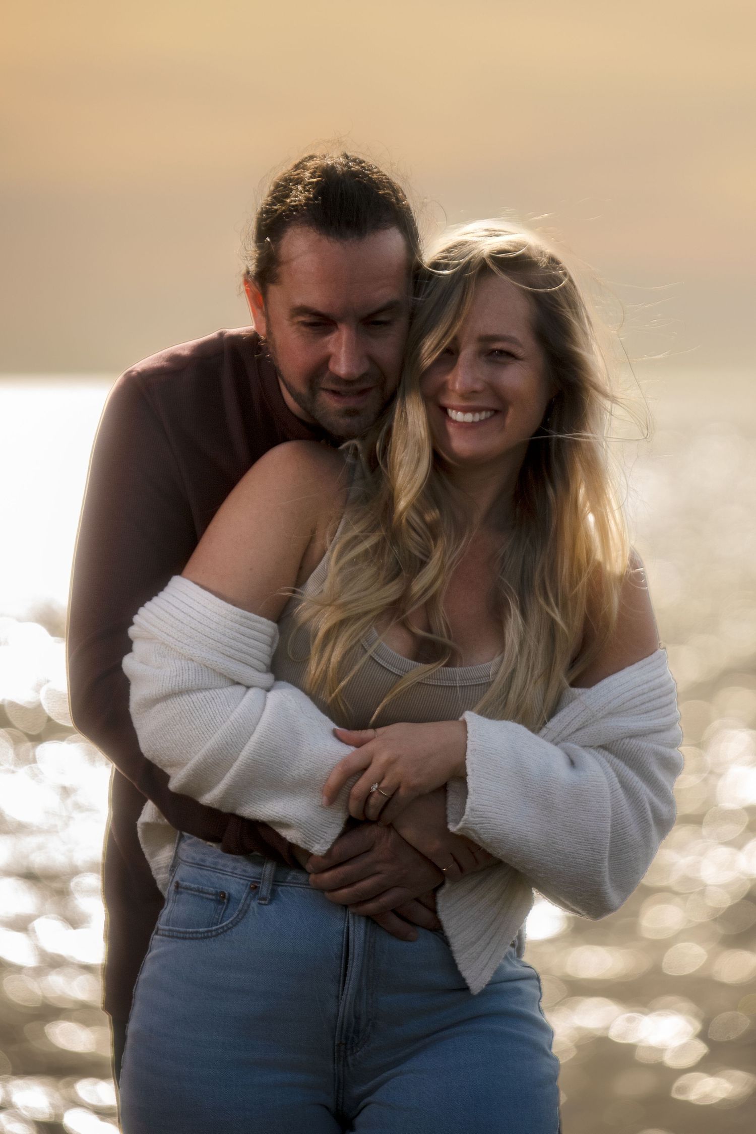 Romantic couple embraces on a beach at sunset with sparkling water in the background.