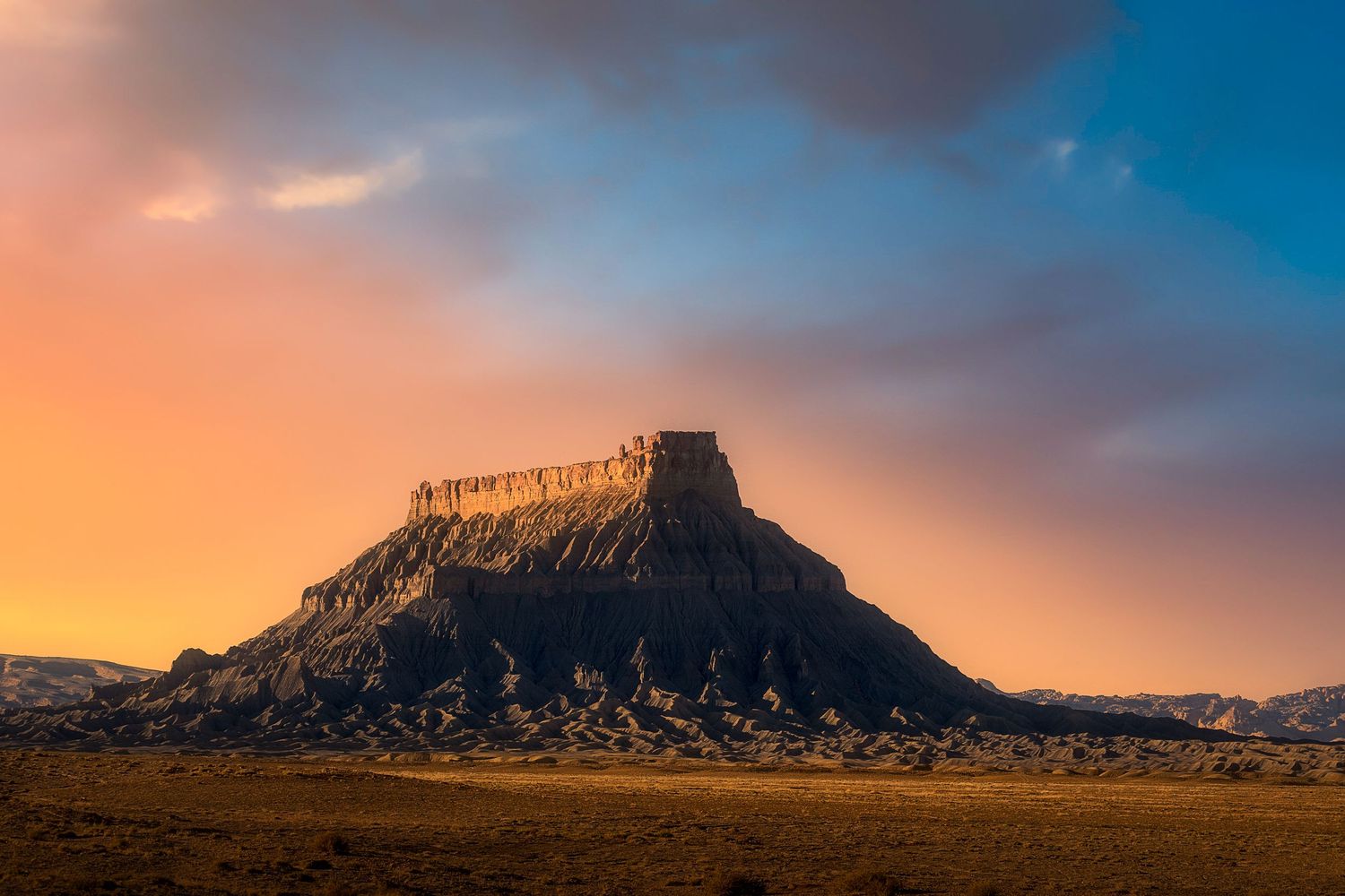 Dramatic sunset over a desert butte with golden light illuminating rocky cliffs against a vivid orange and blue sky.