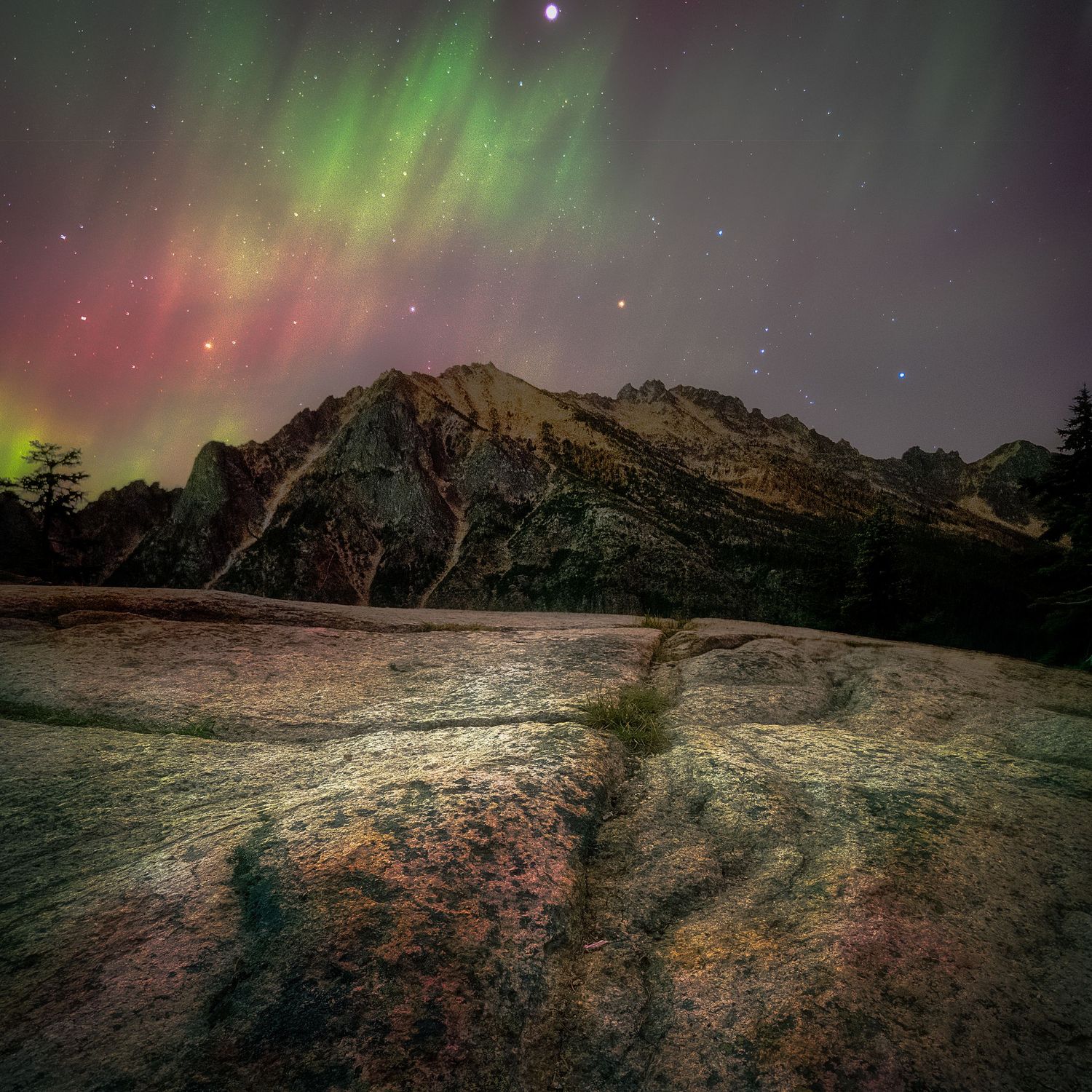 Aurora borealis illuminates a rocky mountain landscape with vivid green and red lights under a starry night sky.