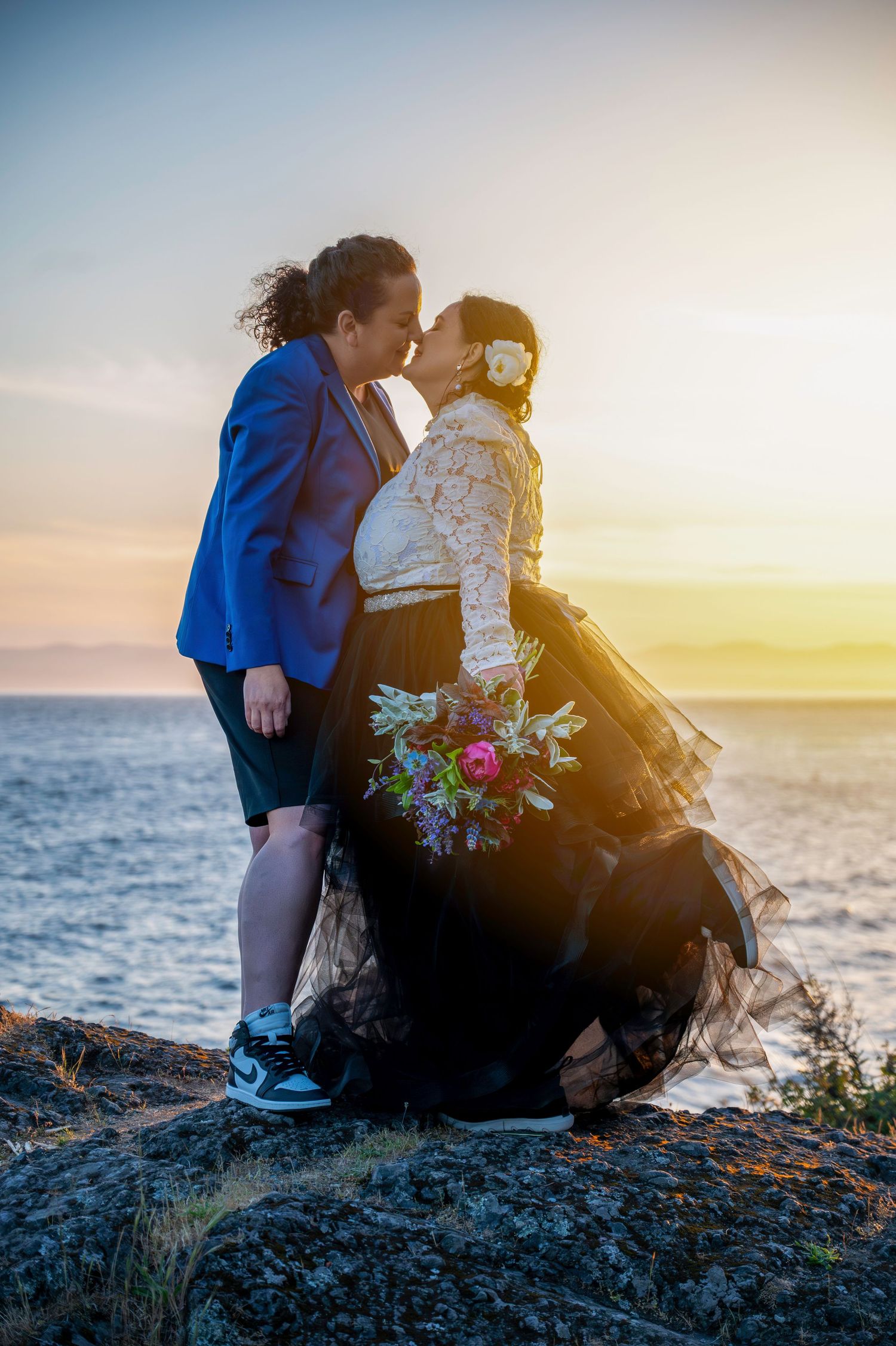 A romantic couple shares a kiss at sunset on a rocky oceanside cliff while holding a colorful bouquet of flowers.