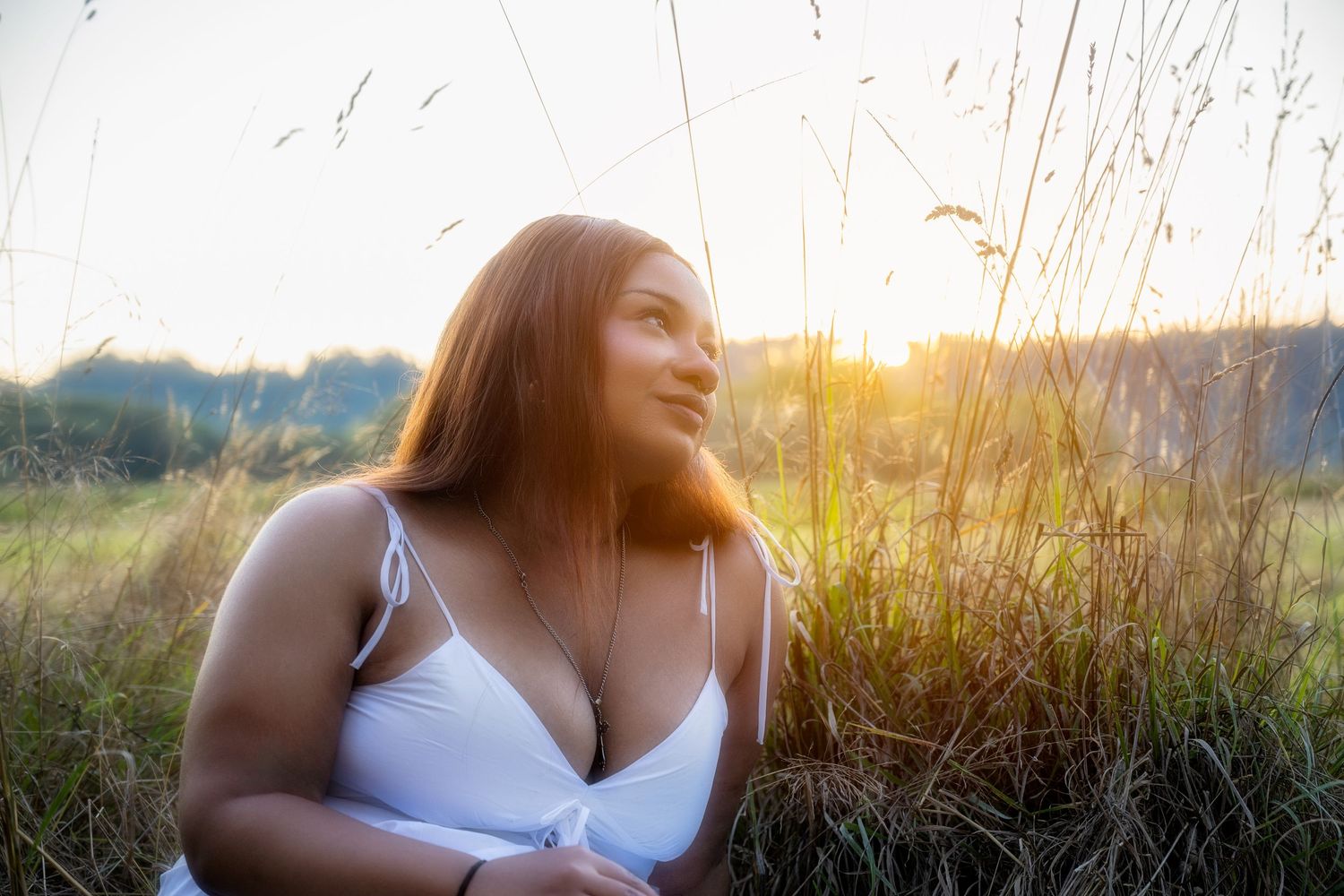 Woman in white dress sitting in tall grass during golden sunset in nature.