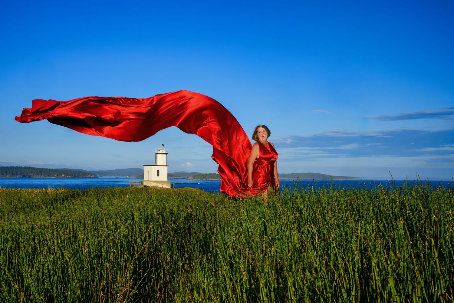 Red fabric billows in the wind against blue sky and green field with white lighthouse in background.