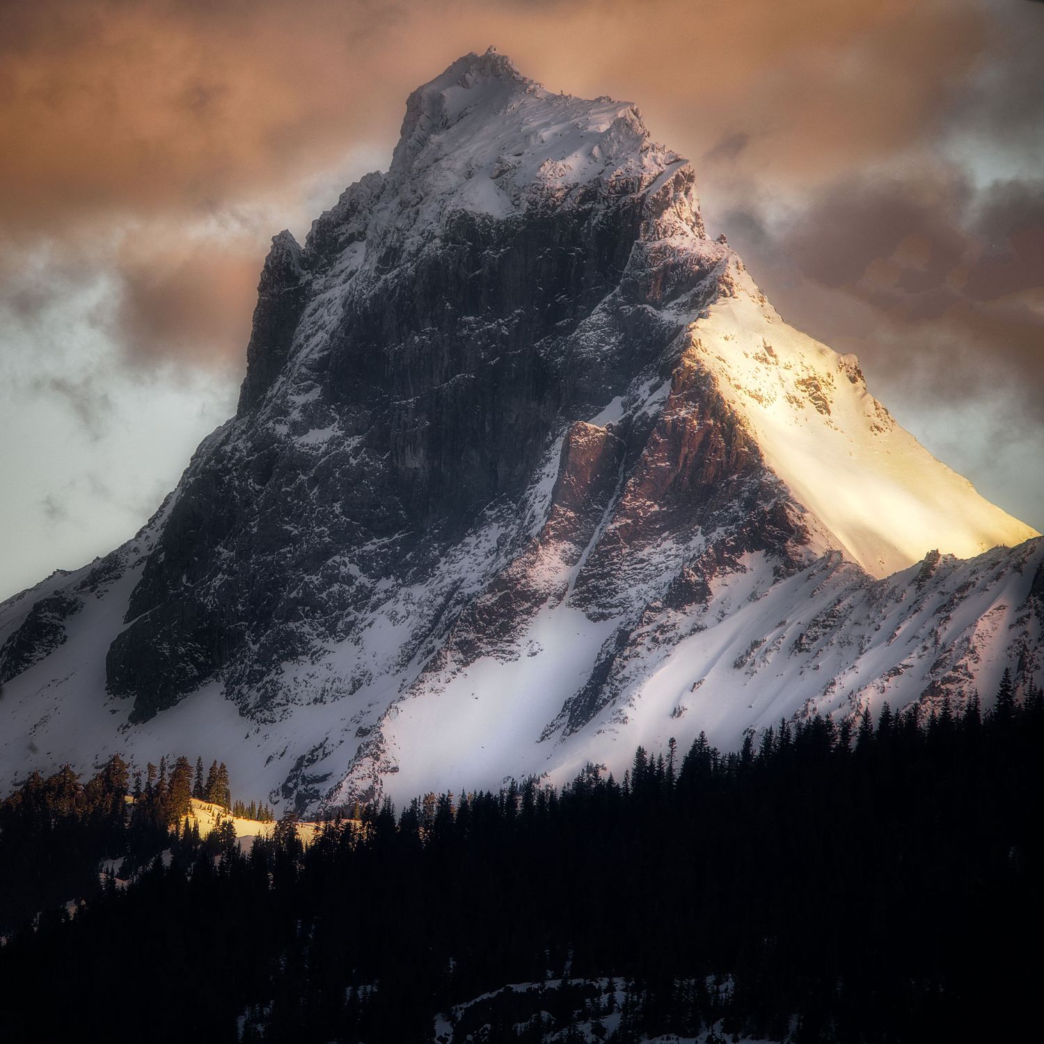 Snow-covered mountain peaks at sunset with dramatic orange clouds and dark silhouetted forest.